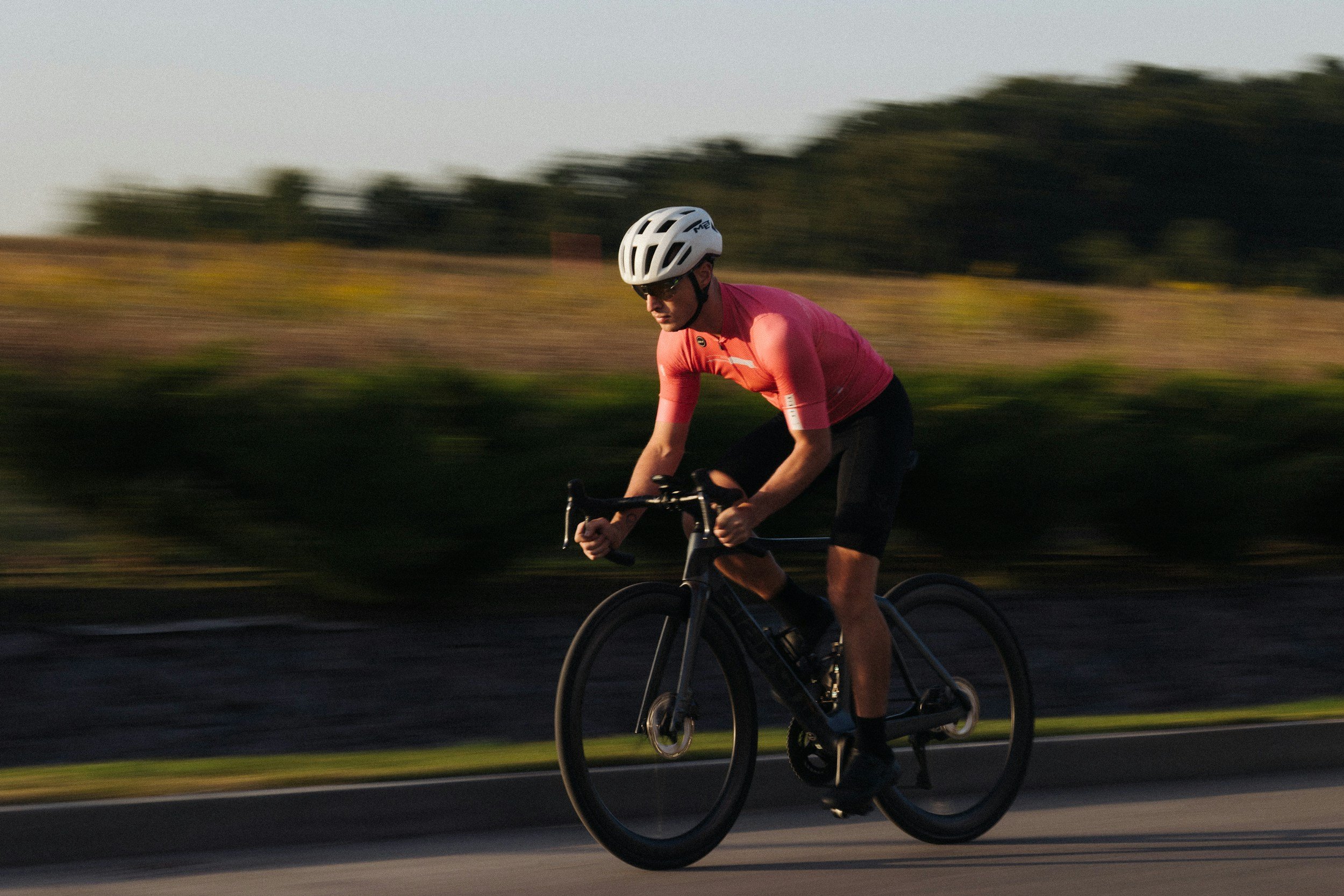 A person wearing a white helmet and a pink shirt riding a black bicycle on a road at sunset.