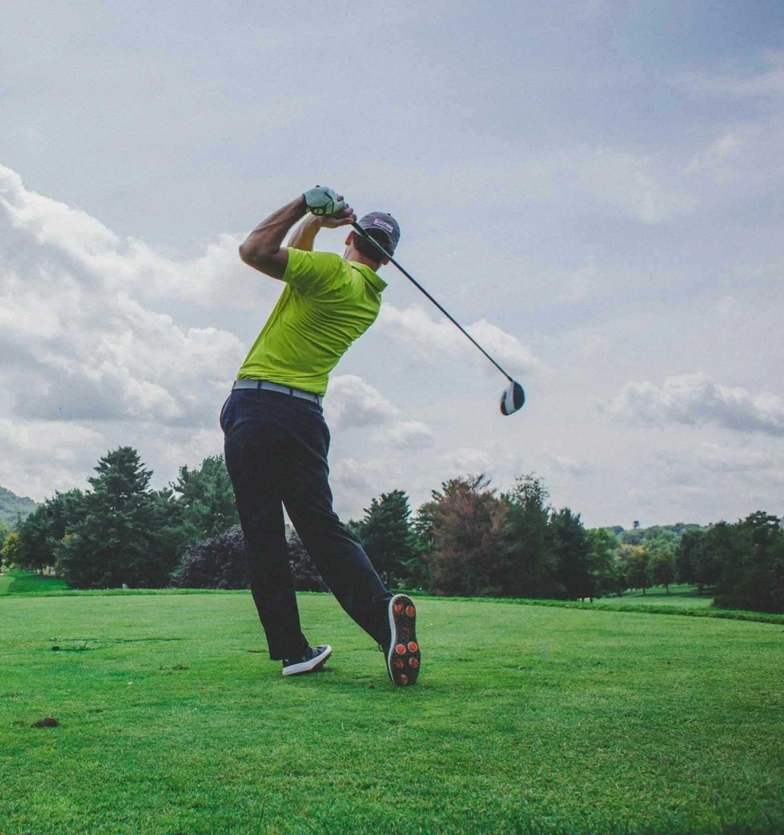 A man in a bright green shirt and black pants swinging a golf club on a golf course under a cloudy sky.