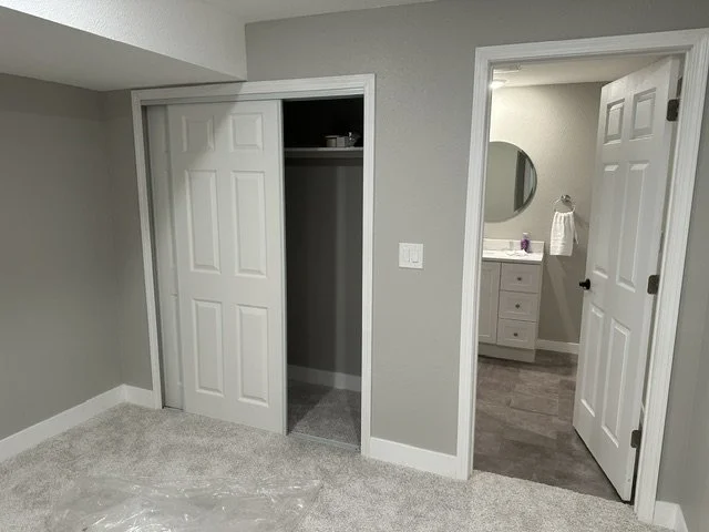 Interior view of a bedroom showing an open closet with sliding doors, a doorway leading to a bathroom with a vanity mirror, sink, and towel, with neutral gray walls and carpeted floor.
