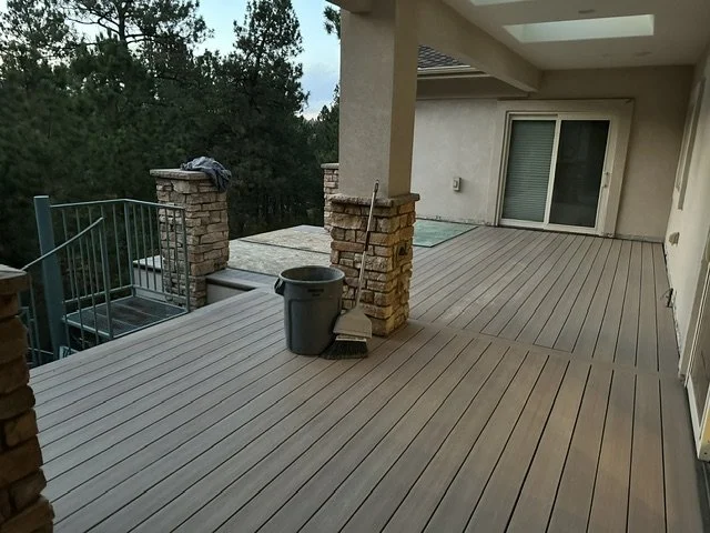 Empty balcony with wooden flooring, brick pillars, a mop and bucket, and sliding glass door leading into house, with trees in the background.