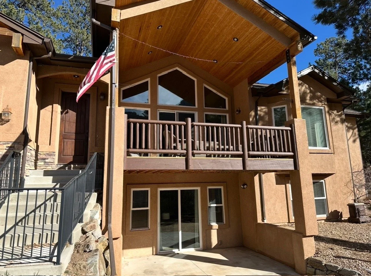 A two-story house with a tan stucco exterior, a wooden balcony with a railing, and large front windows. An American flag is hanging from a flagpole near the front door, which is accessed by stairs with metal railings. The house is surrounded by tall trees and has a gravel yard.