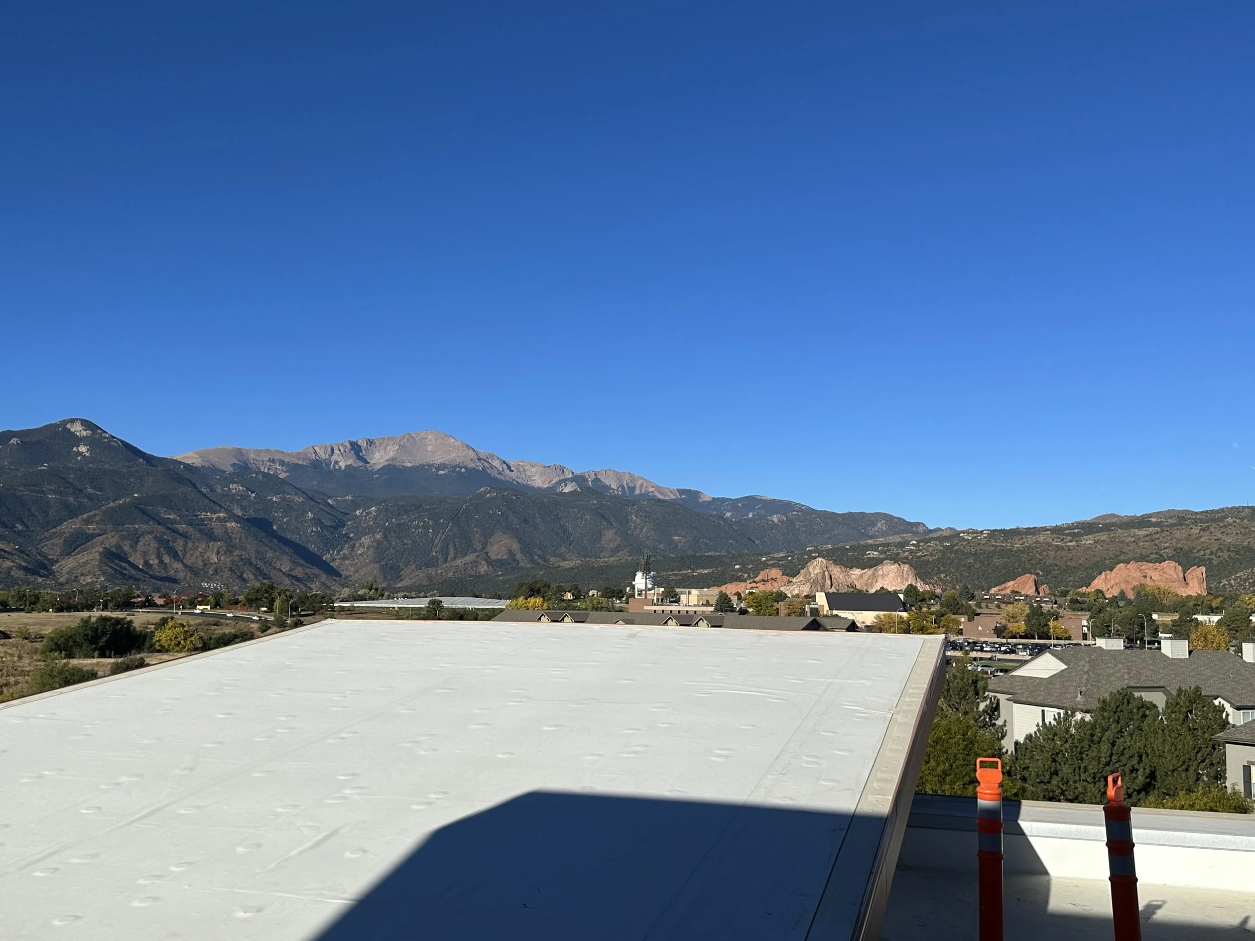 View of distant mountains under a clear blue sky, with a flat roof in the foreground and a few orange traffic barrels visible on the right side.