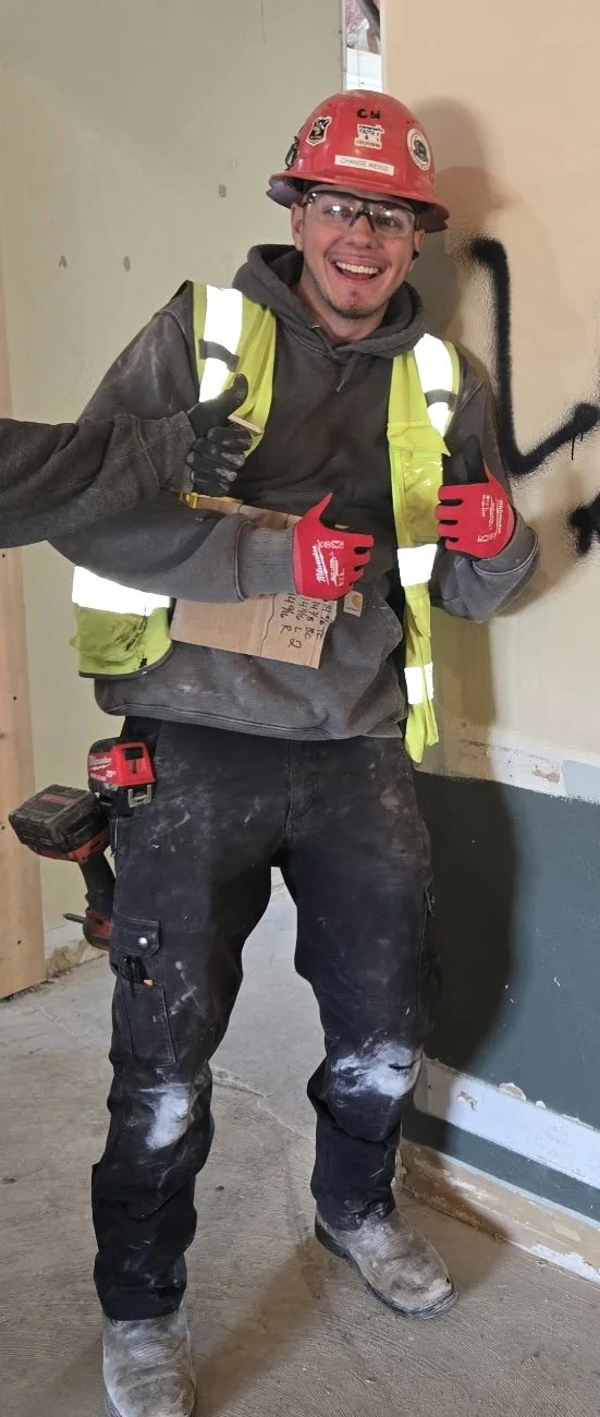 A smiling construction worker in safety gear, including a red helmet, safety glasses, high-visibility vest, and red gloves, giving a thumbs-up while standing indoors on a construction site.