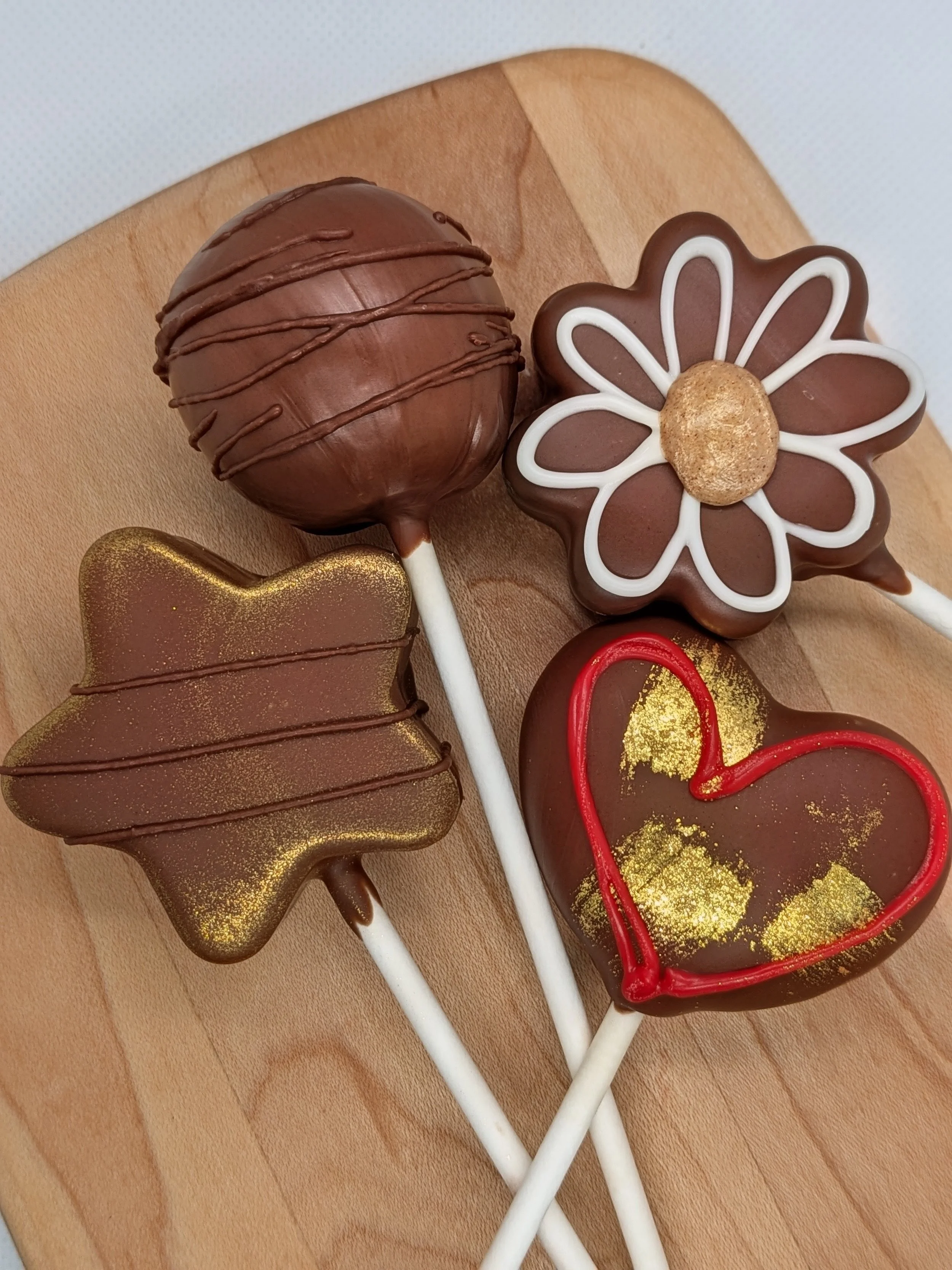 Decorative chocolate cake pops shaped like a heart, flower, and other designs, with red and gold accents, on a wooden surface.