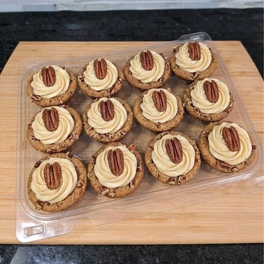 Twelve light brown cookie cups with pecan chunks, topped with a buttercream swirl and a whole pecan displayed in a plastic clamshell tray on a wooden board on a kitchen counter. and