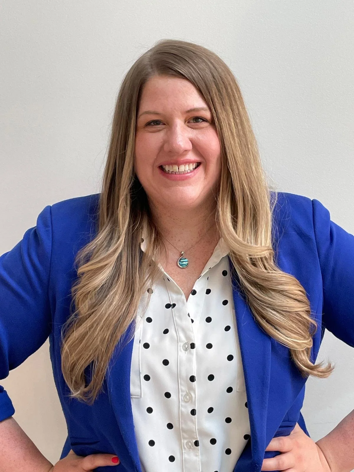 A woman with long, wavy hair smiling, wearing a blue blazer over a white blouse with black polka dots, and a necklace with a pendant, standing against a plain white wall.