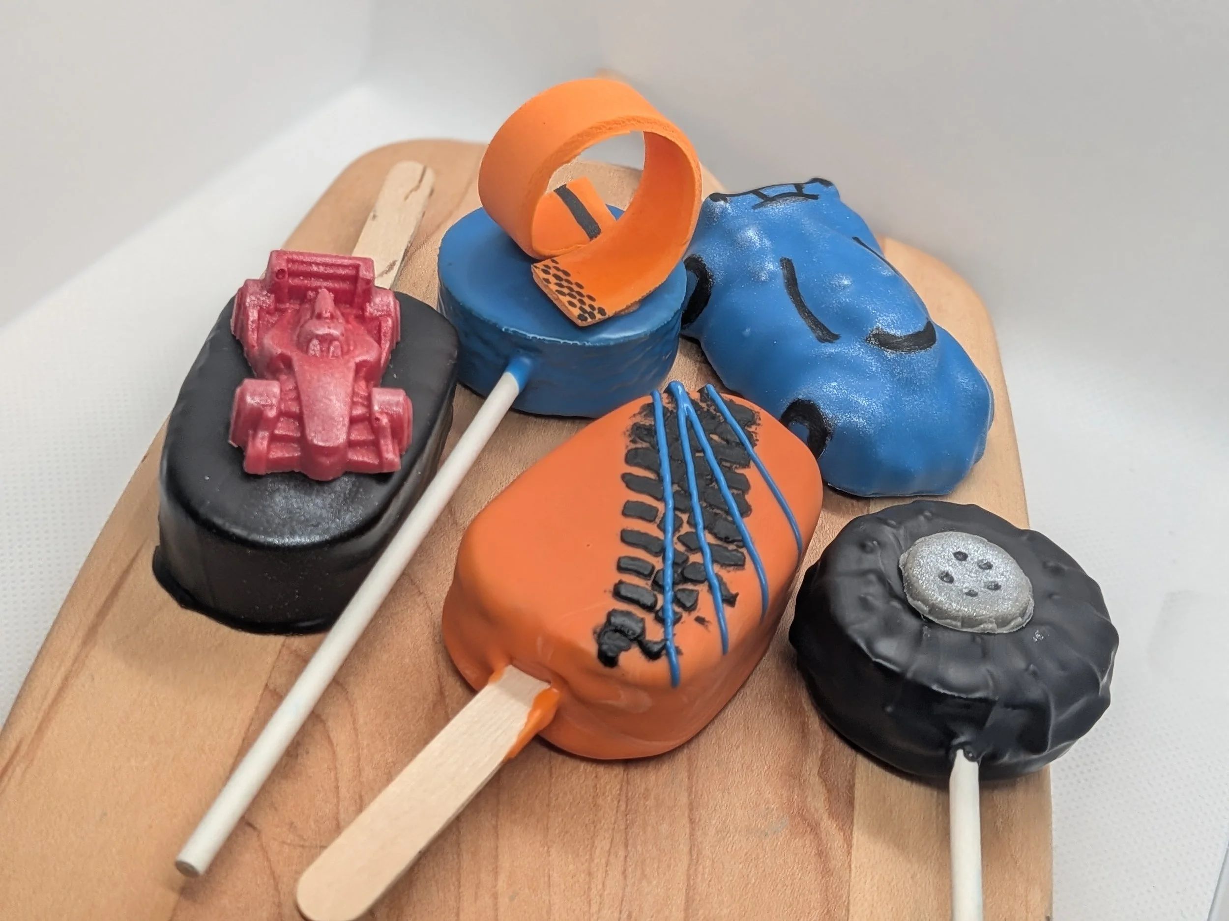 Decorative cake pops and cakesicles in orange, blue, black, and red colors shaped like a race car, a tire, and a racing track loop on a wooden board.