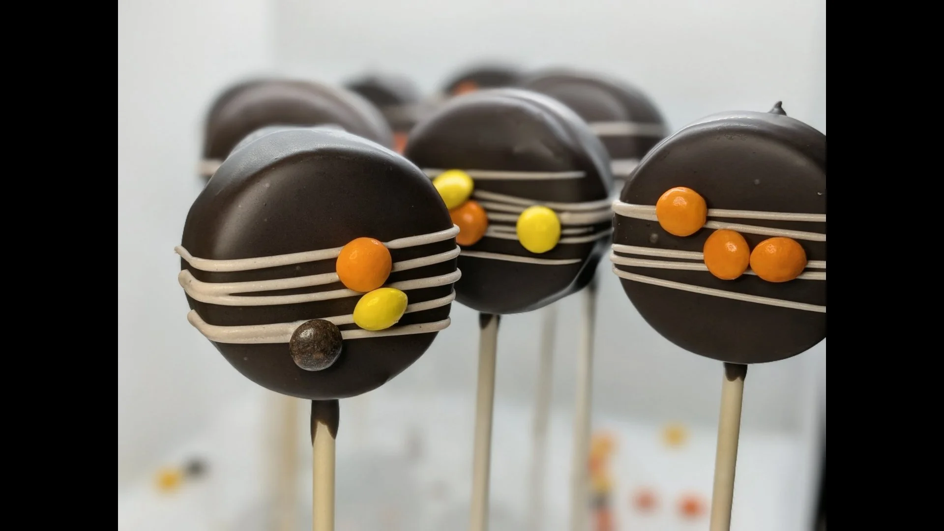 Close-up of spherical chocolate cake pops decorated with Reese's Pieces and white icing lines, mounted on sticks against a plain background.