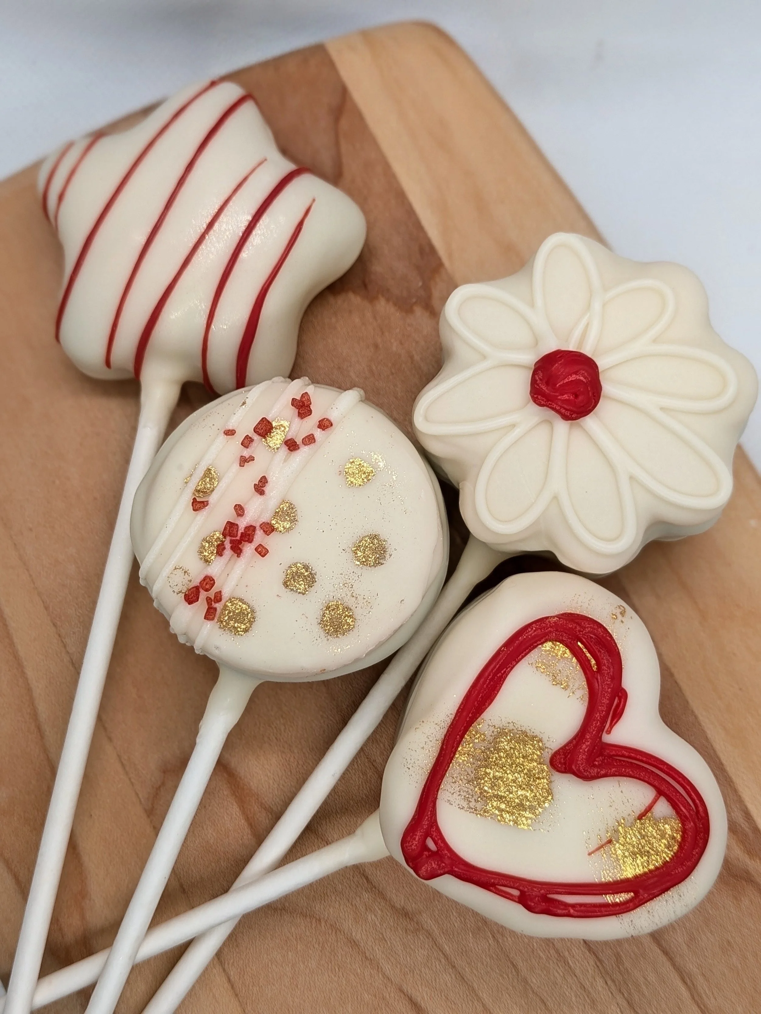 Four decorated cake pops with white chocolate coating, red and gold sprinkles, and red icing in a heart shape, arranged on a wooden surface.