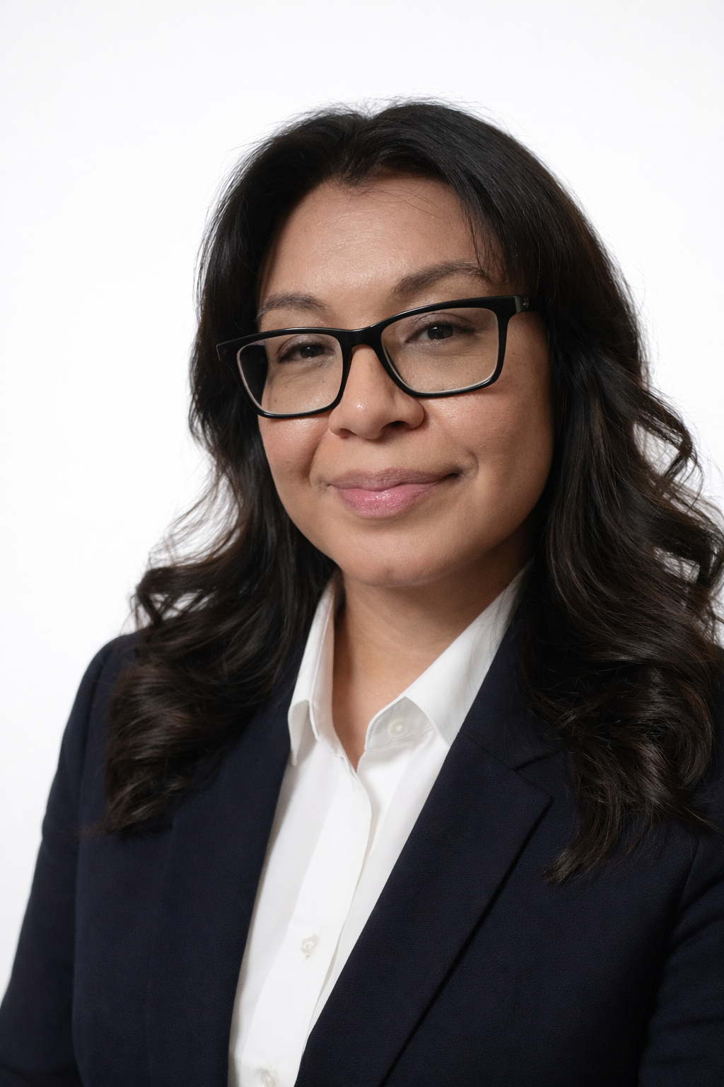 Portrait of a woman with black hair wearing glasses, a white collared shirt, and a black blazer, smiling at the camera against a plain white background.