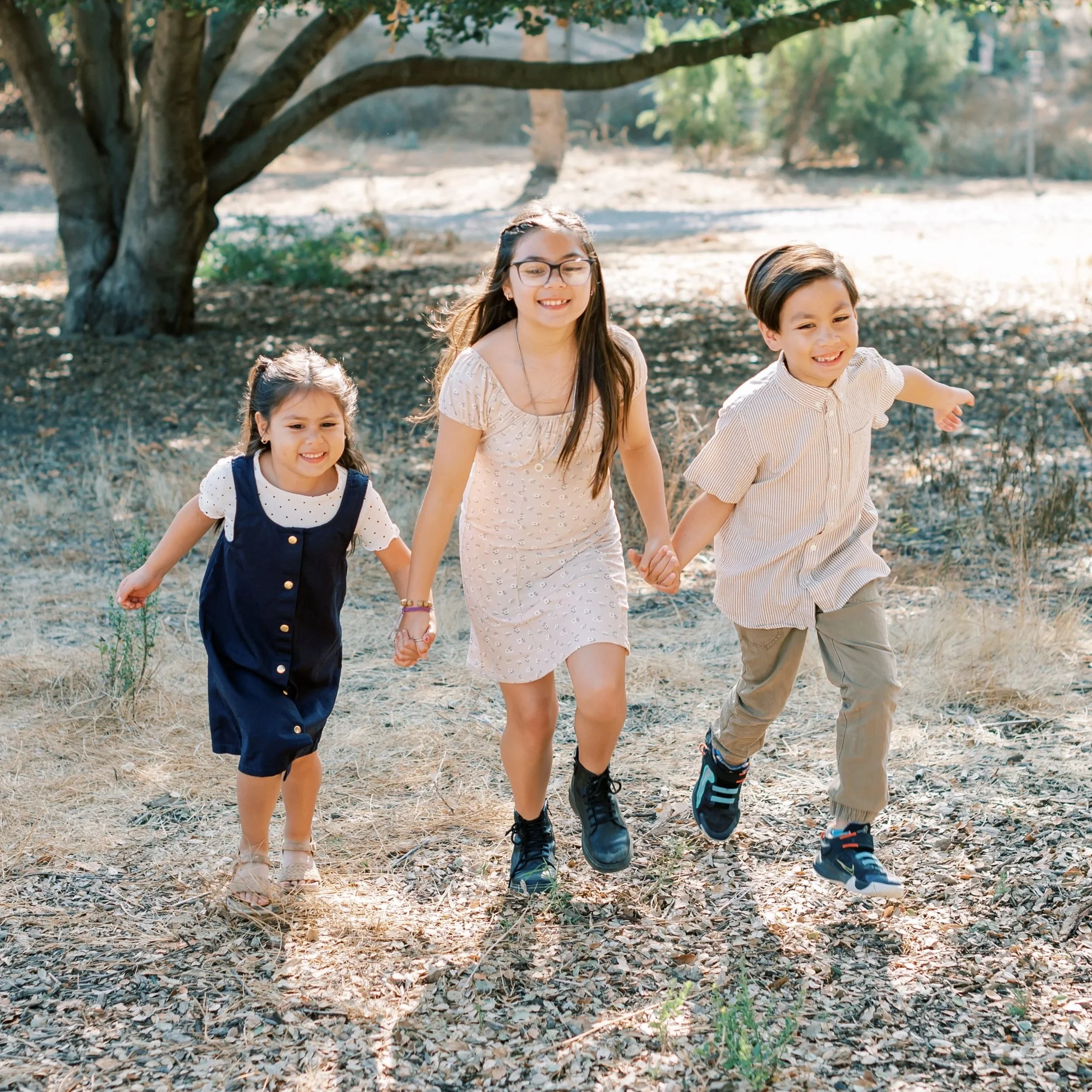 Three children walking hand in hand outdoors on a dirt path under a tree in a sunny park.