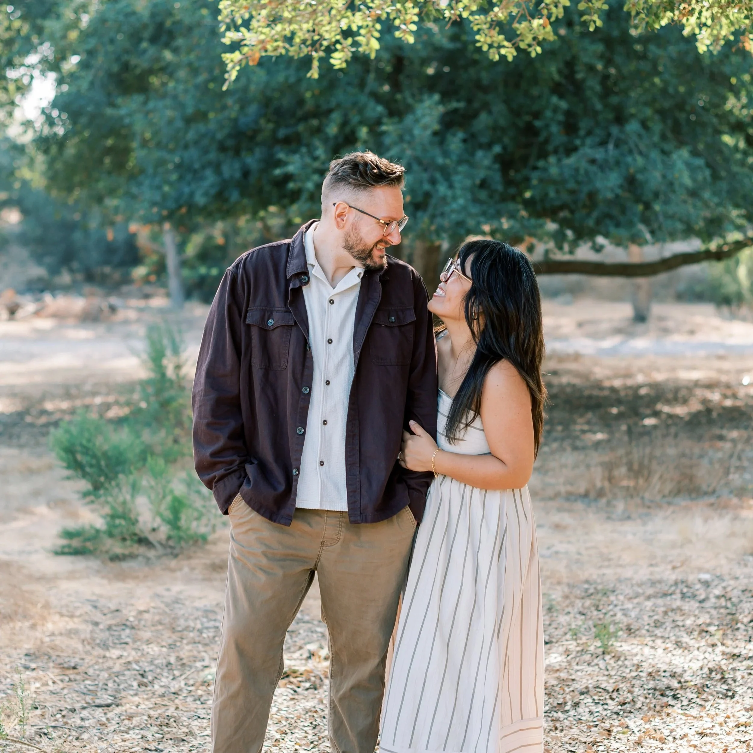 A man and woman standing close together outdoors, smiling and looking at each other. The man has short hair, glasses, a beard, and is wearing a dark jacket and beige pants. The woman has long dark hair, glasses, and is dressed in a sleeveless white dress with black vertical stripes. They are surrounded by trees and greenery.
