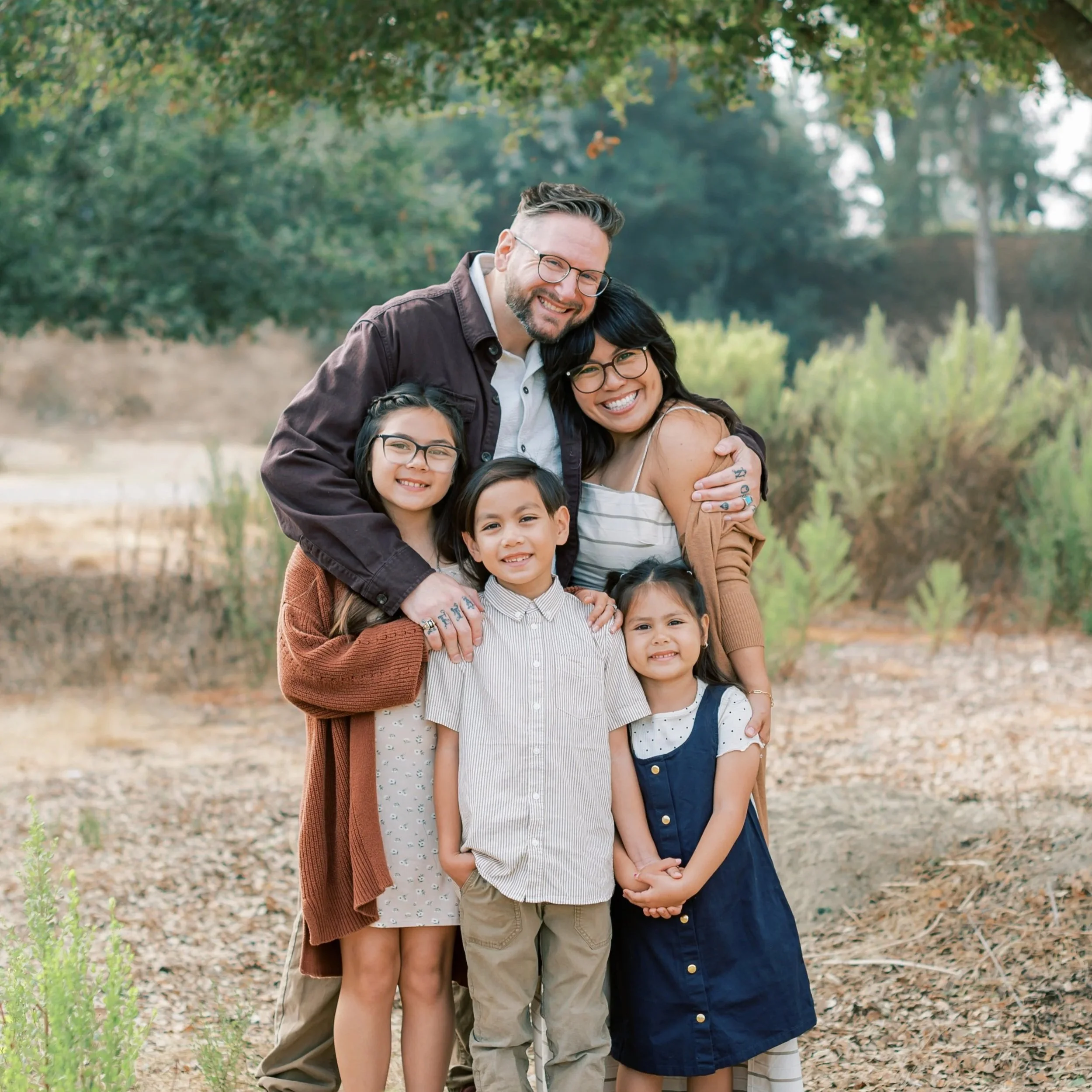 Family smiling and hugging outdoors in a park with green trees in the background.