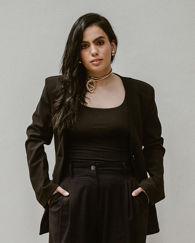 A woman with long dark hair wearing a black blazer, black top, and black pants, standing against a plain wall.
