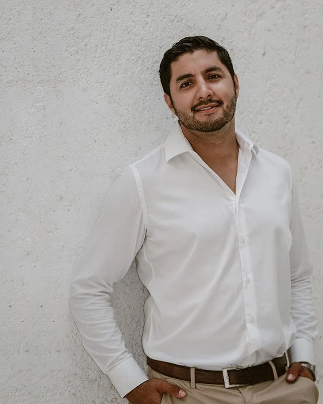 A man with dark hair and a beard wearing a white dress shirt and beige pants leaning against a textured off-white wall.
