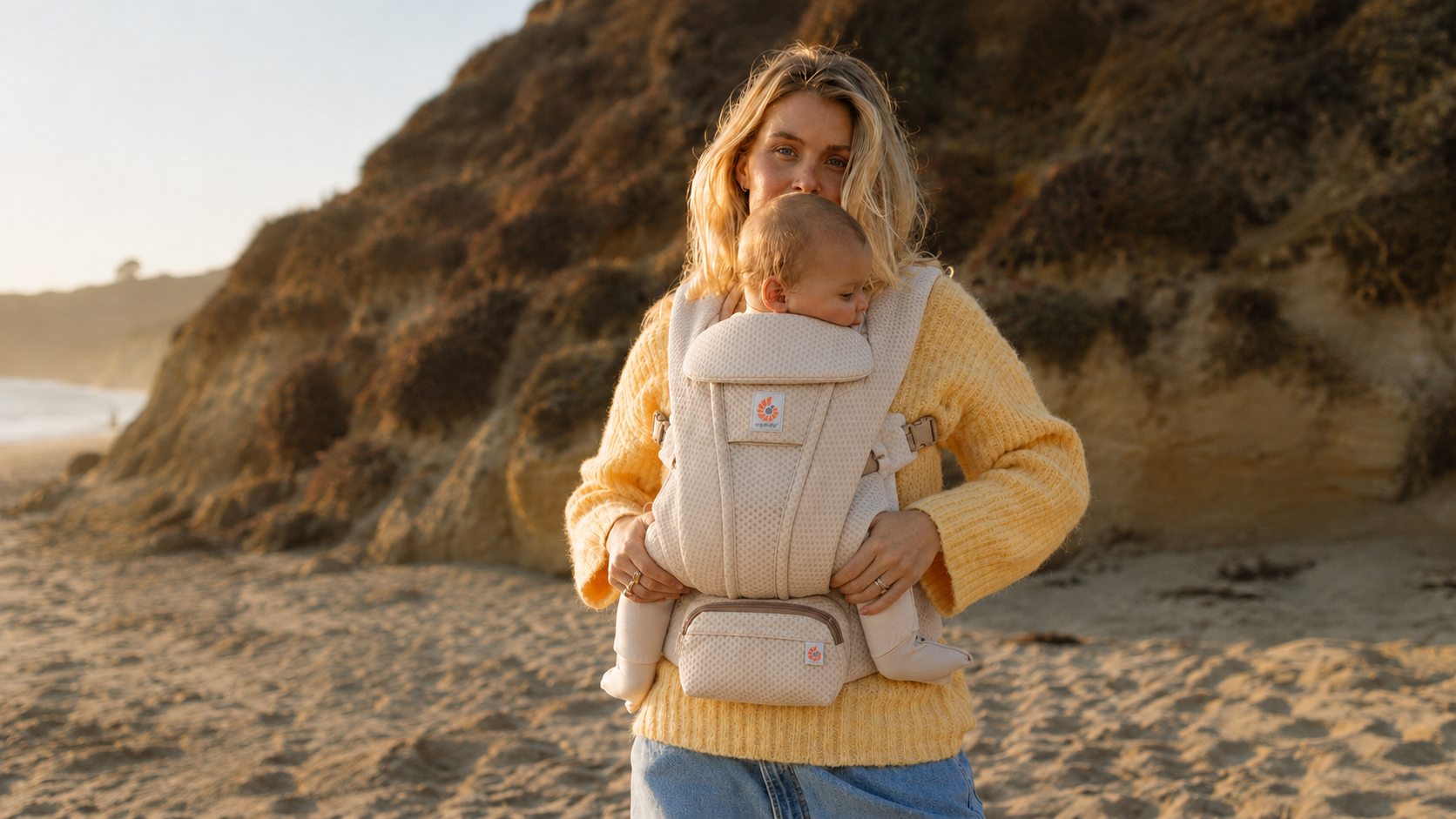 A woman carrying a young child in a beige baby carrier on a beach at sunset, with a rocky hillside in the background.