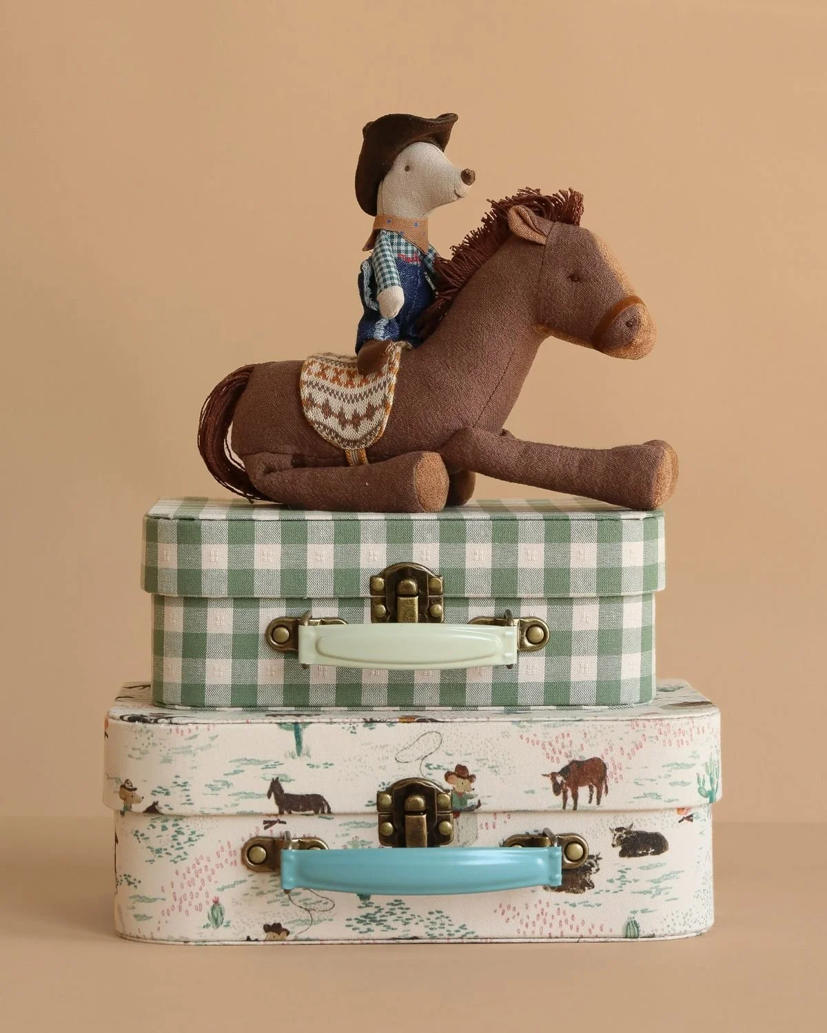 A plush toy of a boy wearing a cowboy hat and vest, riding a stuffed brown horse, placed on stacked decorative boxes with animal and nature prints.