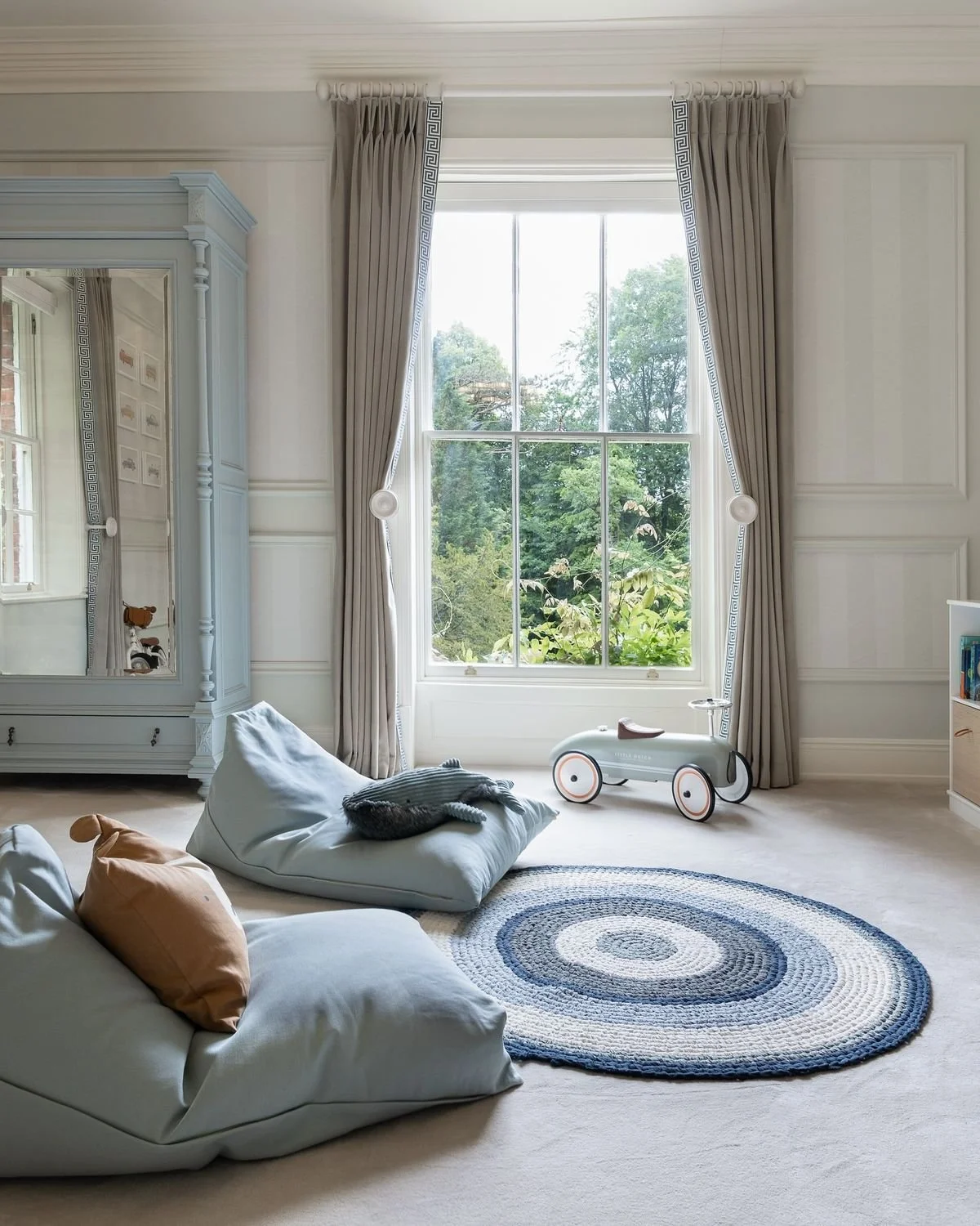 A bright, cozy child's bedroom with large window, beige curtains, white walls, round rug, stuffed animal on floor cushion, toy ride-on car, and white furniture.
