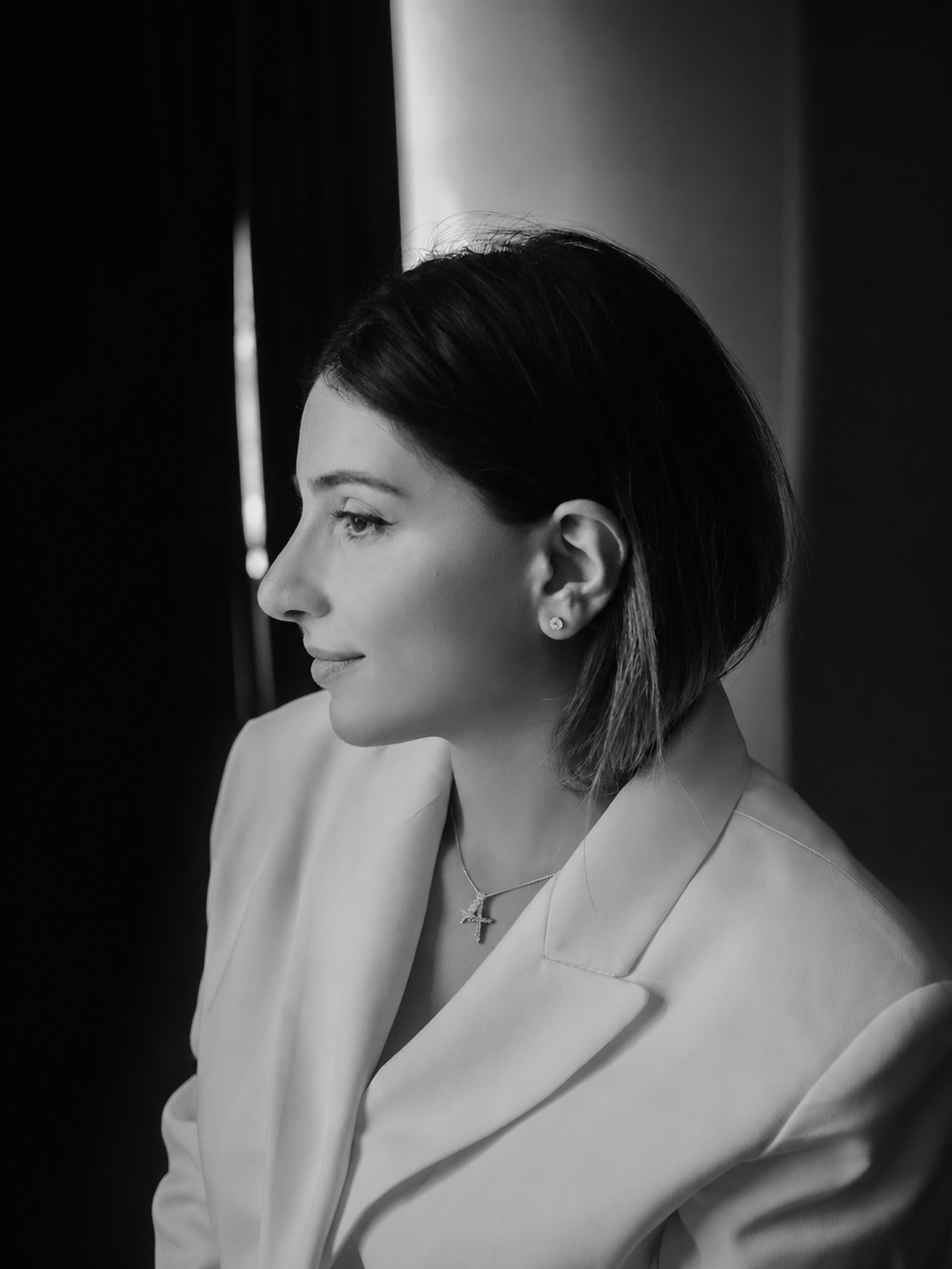 A woman with short dark hair and earrings, wearing a light-colored blazer and a cross pendant necklace, looking to the side in a black and white profile portrait.