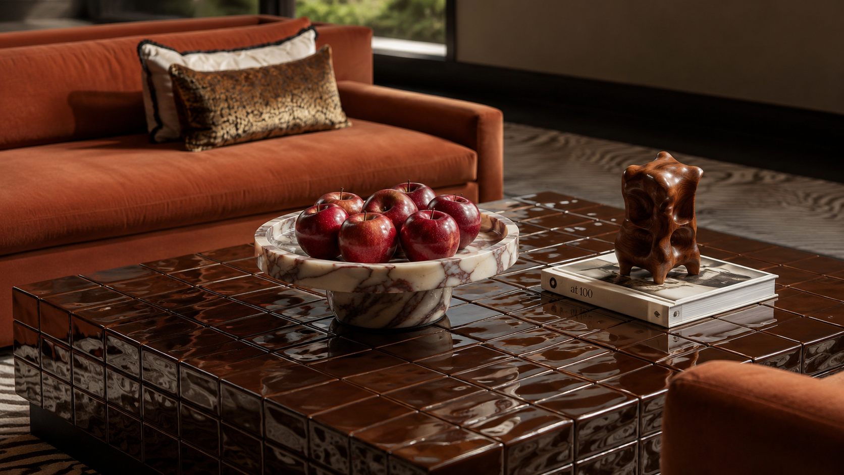 Living room with an orange sofa, patterned pillows, a reflective tiled coffee table, a marble bowl with red apples, and a decorative wooden sculpture.