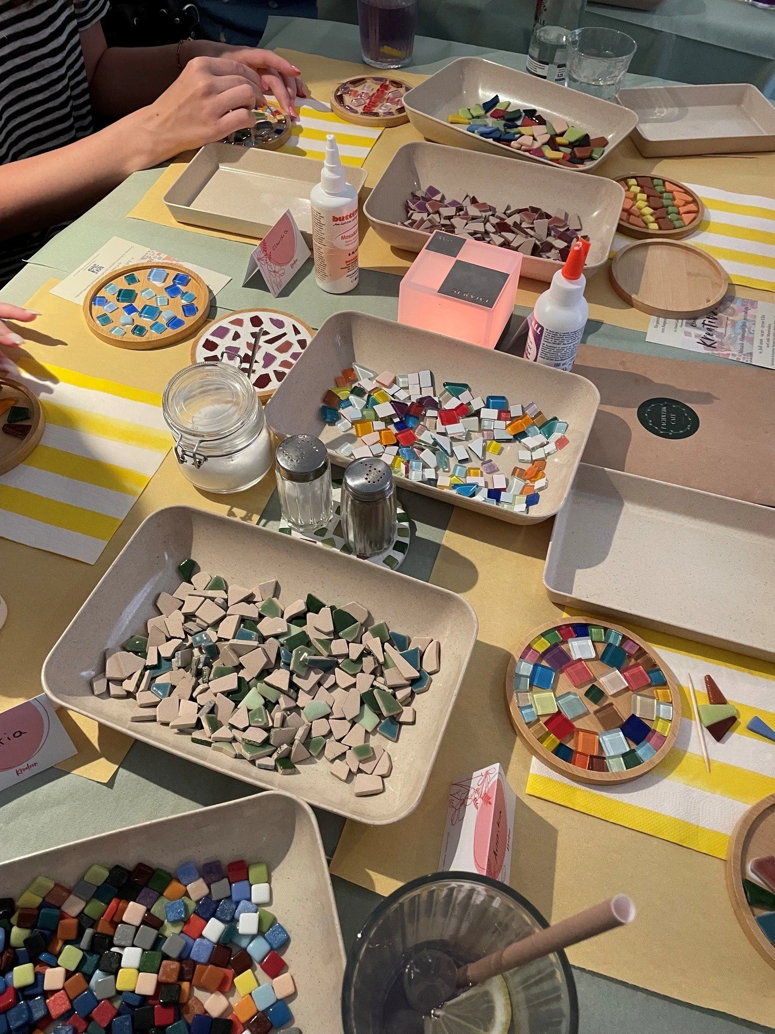 Mosaiksteine in verschiedenen Farben und Formen auf einem Tisch, mit Bastelmaterialien und Getränken.