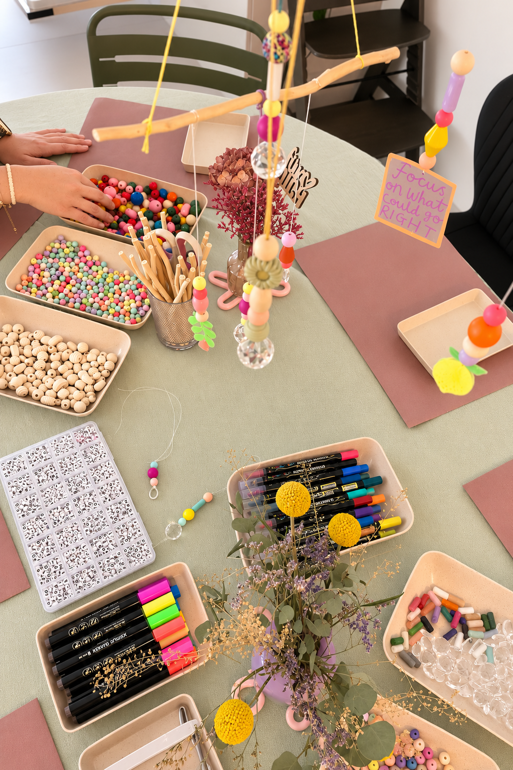 Bunte Bastelmaterialien auf einem Tisch, inklusive Perlen, Marker, Holzstäbchen, und Dekorationen, mit einer kleinen Pflanze in der Mitte.