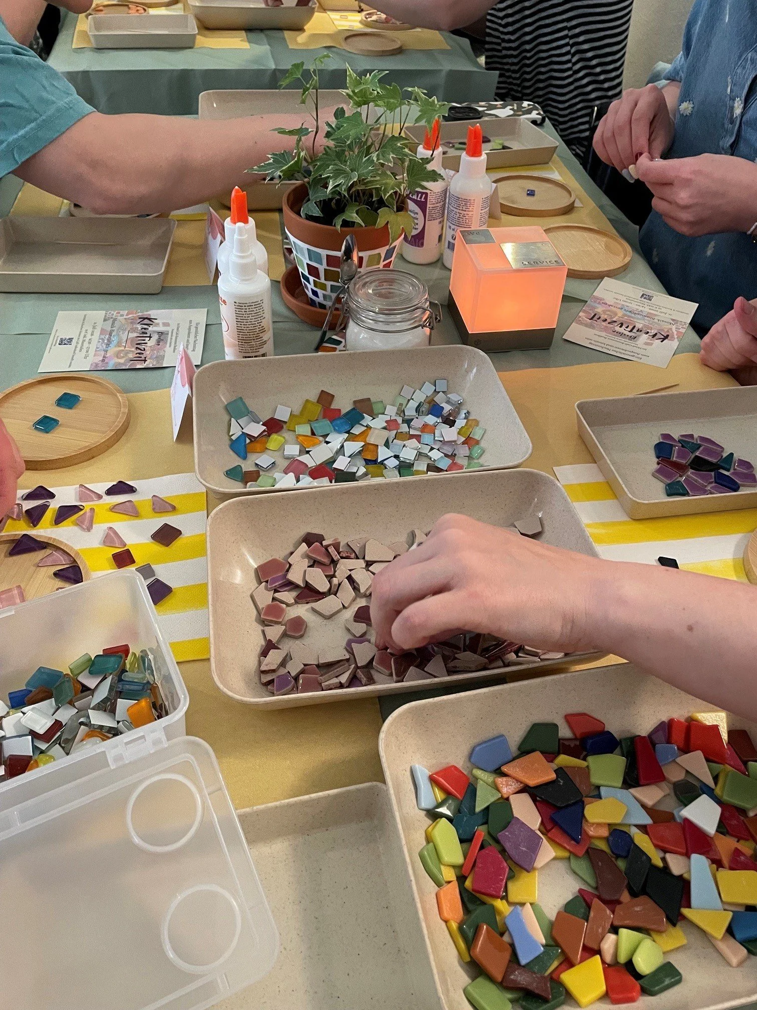 Eine Gruppe von Menschen, die an einem Tisch mit Mosaiksteinchen arbeitet. Es gibt Schalen mit verschiedenen bunten Steinchen, Kleber und andere Bastelmaterialien auf dem Tisch. Im Vordergrund sind verschiedene farbige Steinchen sichtbar, die für ein Mosaikprojekt vorbereitet werden.