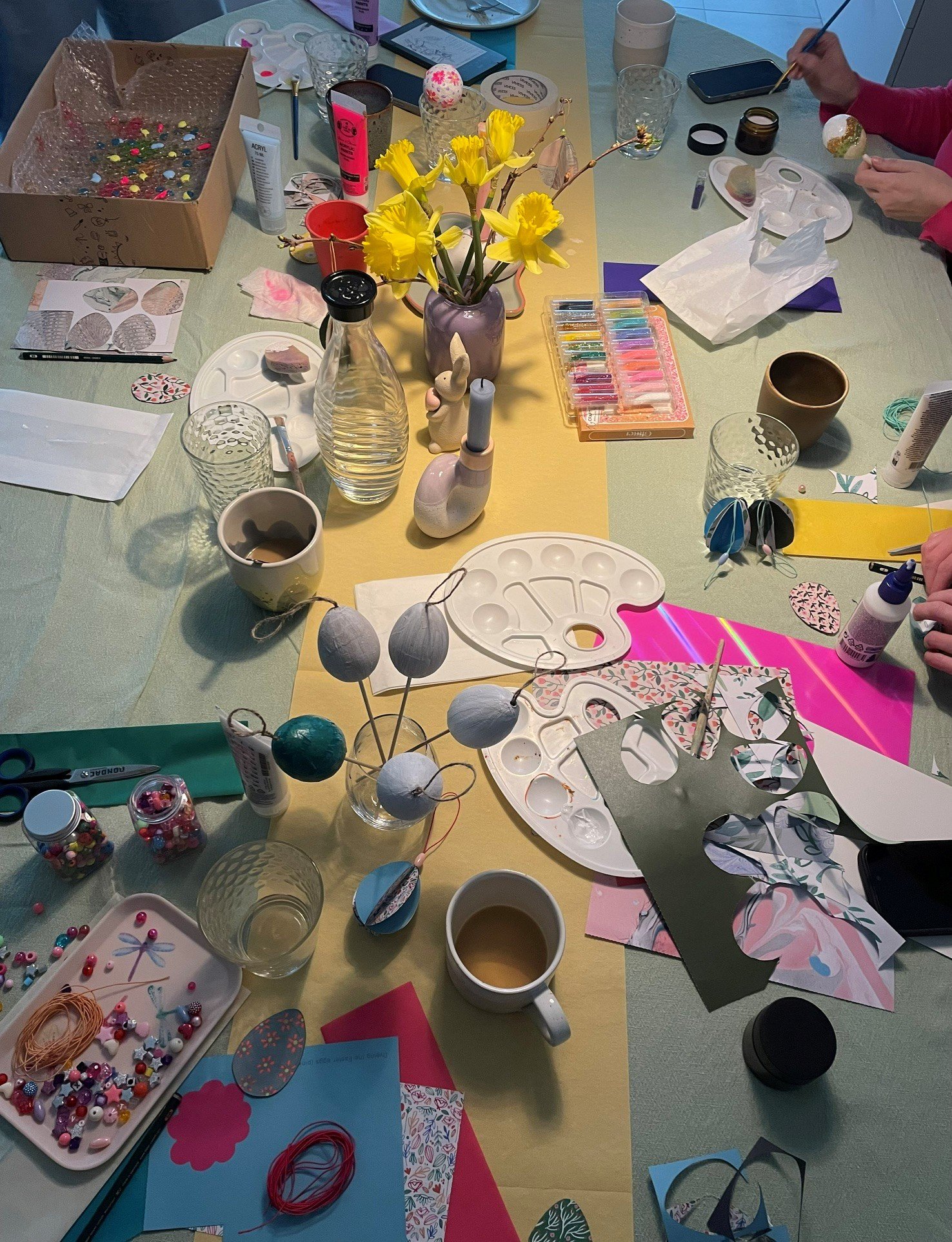 Der Tisch ist mit Bastelmaterialien bedeckt, darunter bunte Perlen, Papier, Messer, Schere, Farben, Kerzen, Blumen und kreative Bastelwerkzeuge. Es scheint eine Bastel- oder Kunstaktivität stattzufinden.