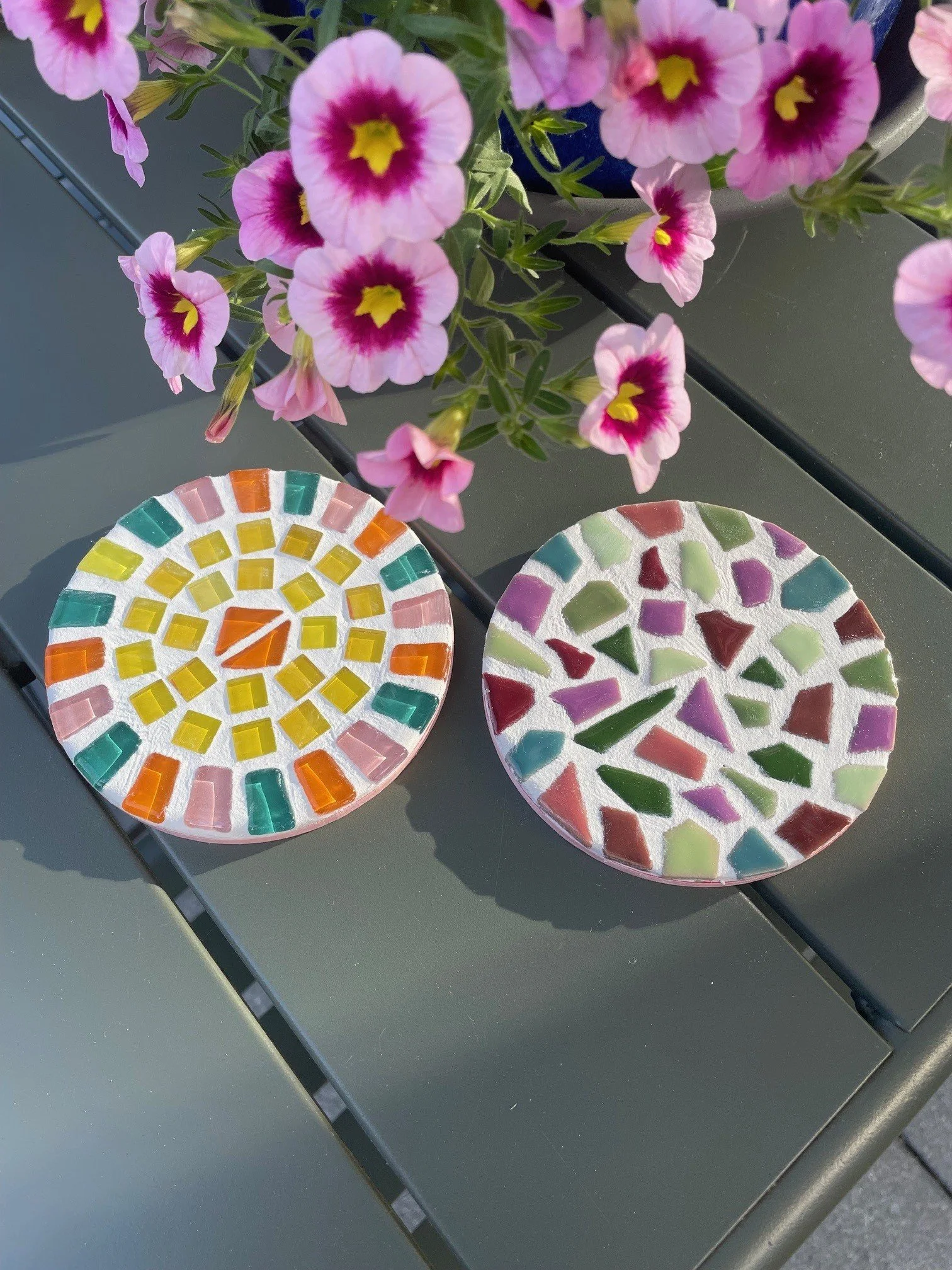 Bunte Mosaikuntersetzer neben pinken Petunien in einem Blumentopf auf einem grauen Tisch.