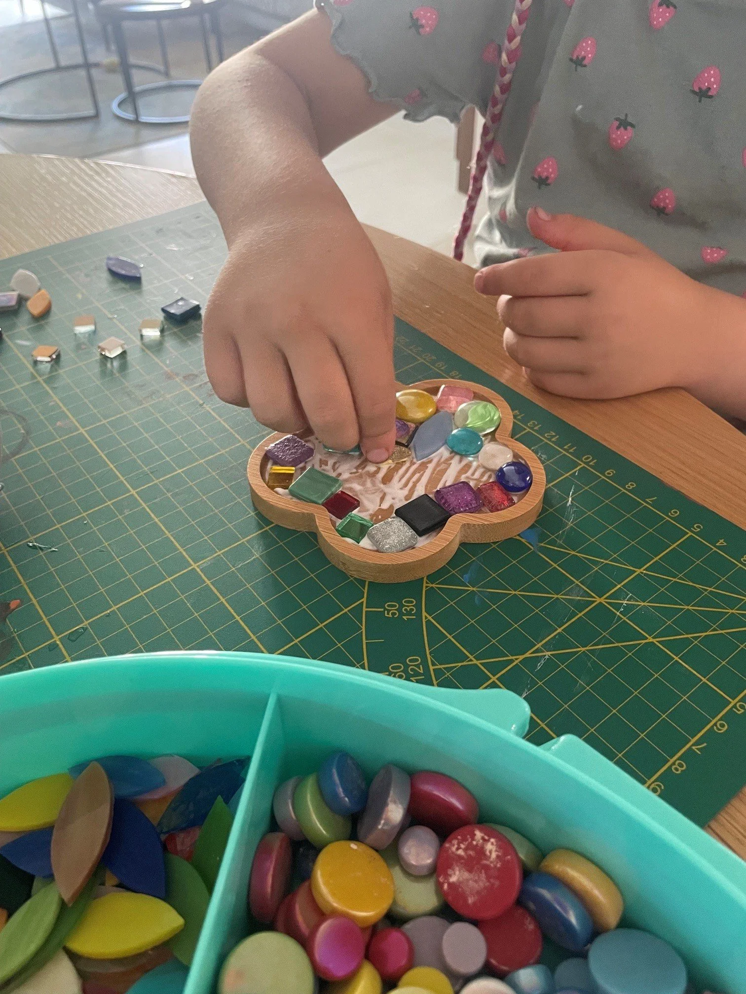 Ein Kind sortiert bunte Holzknöpfe auf einem Schneidebrett. Im Vordergrund eine blaue Schüssel mit weiteren bunten Knöpfen, im Hintergrund eine Grünfläche mit einem Bastelset. Das Kind trägt ein graues Oberteil mit kleinen Erdbeeren.