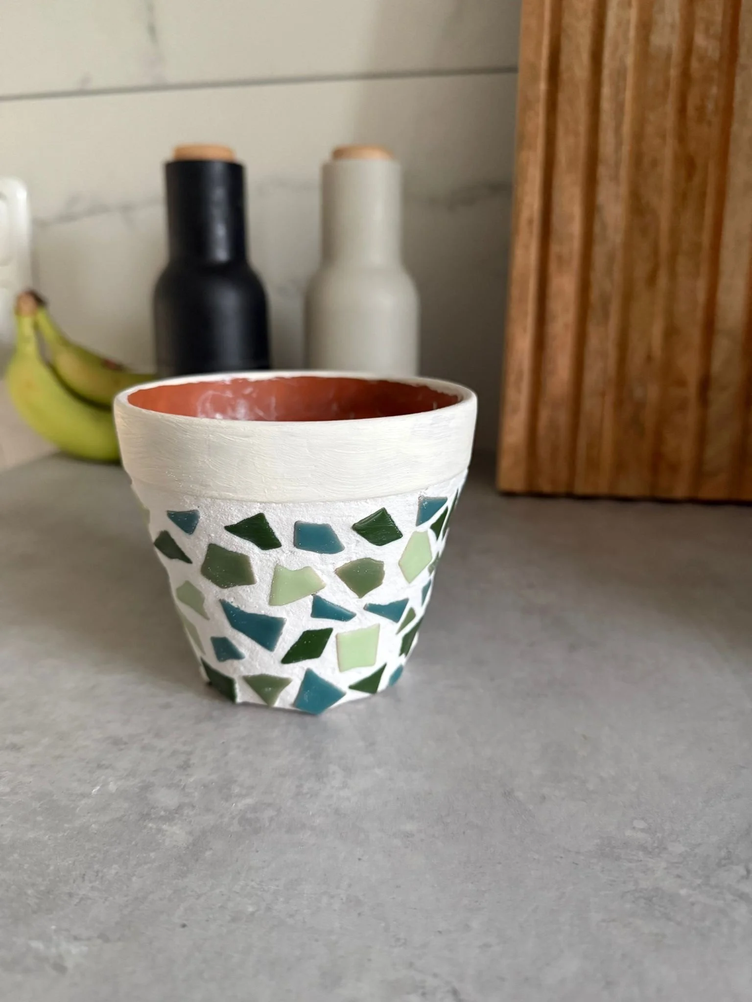 Blumentopf aus Terrakotta gestaltet mit Mosaiksteinen auf einer grauen Oberfläche, hinter zwei Flaschen, einer schwarzen und einer weißen, und Bananen.