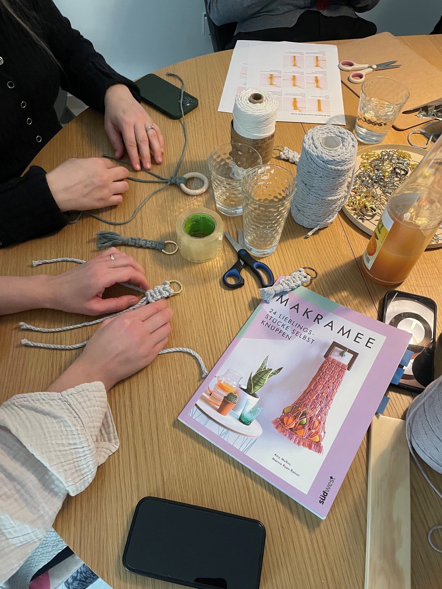 Menschen arbeiten an Makramee-Projekt an einem Tisch mit Makramee-Anleitung, Garnrollen, Scheren, Glasschüsseln und Schmuckteilen.