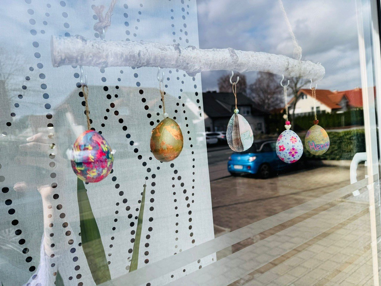 Bunte Osterdekoration aus bemalten Eiern, die an einem Ast hängen, im Fenster mit reflektiertem Hintergrund von Häusern und einem Auto