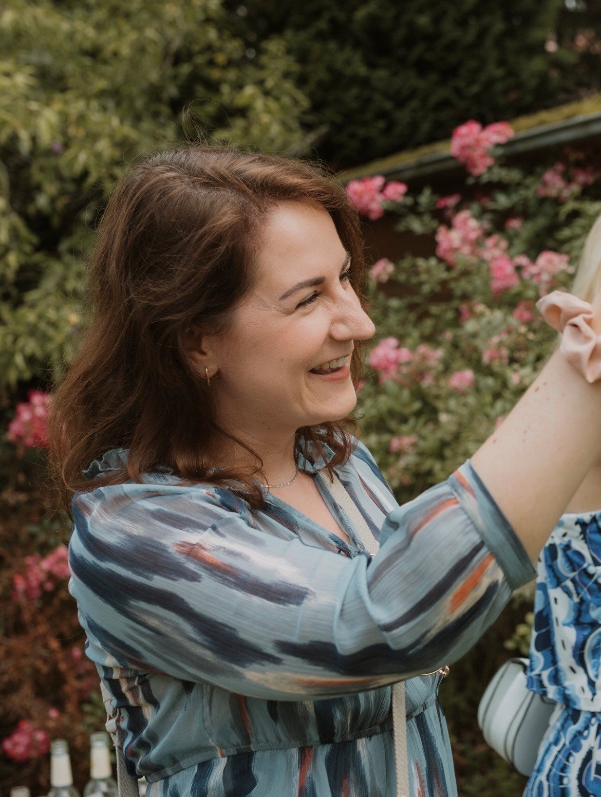 Eine lächelnde Frau mit braunen Haaren, trägt ein blaugemustertes Kleid, steht im Garten mit rosa Blumen im Hintergrund.