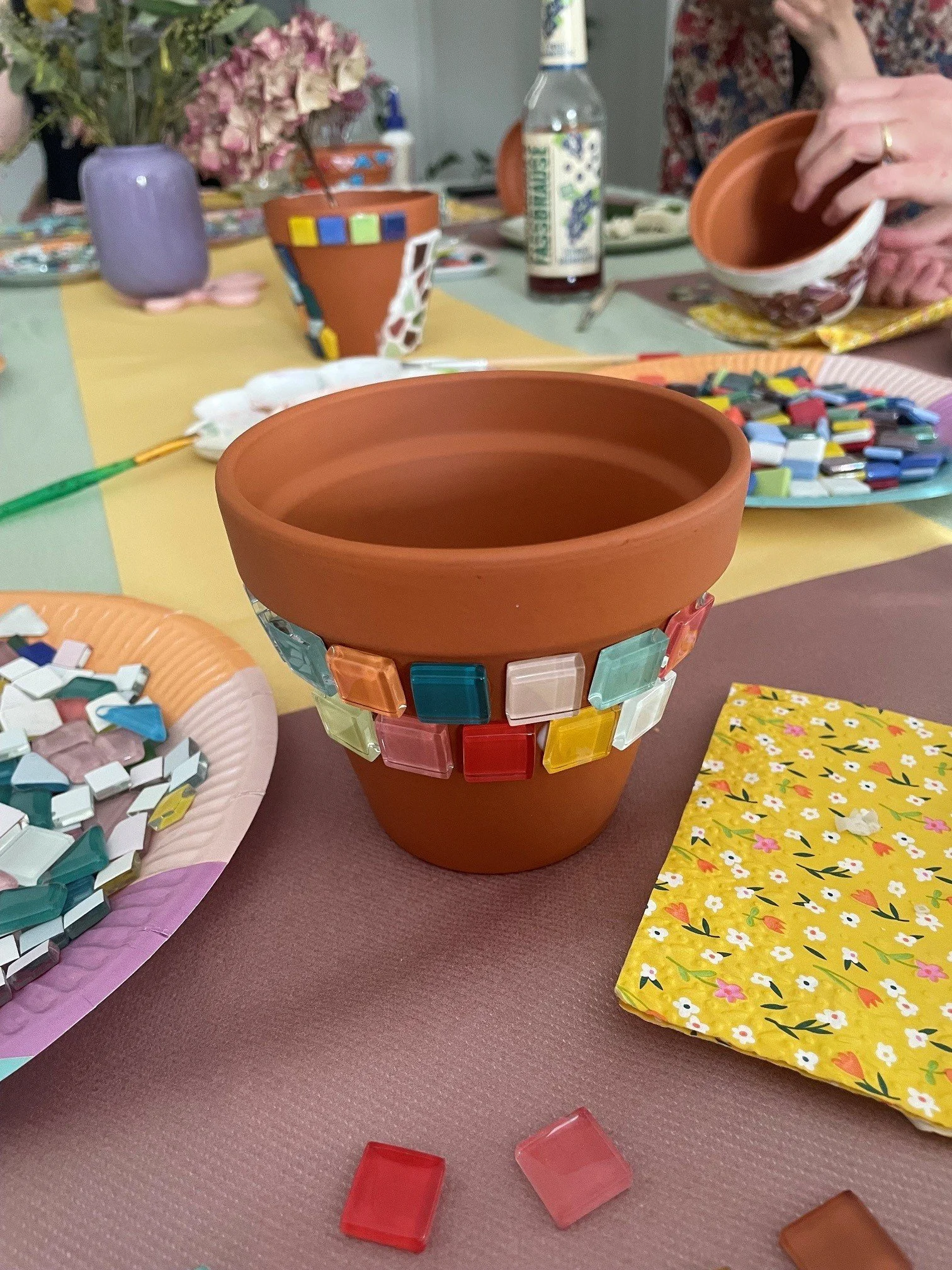 Ein bemalter Terrakottatopf mit bunten quadratischen Fliesen um die Oberseite auf einem Tisch, im Hintergrund sind Blumen, eine Flasche, eine Tasse und ein Pappteller mit Müsli oder Cornflakes zu sehen, die Szene zeigt eine Kaffeepause oder ein Treffen.