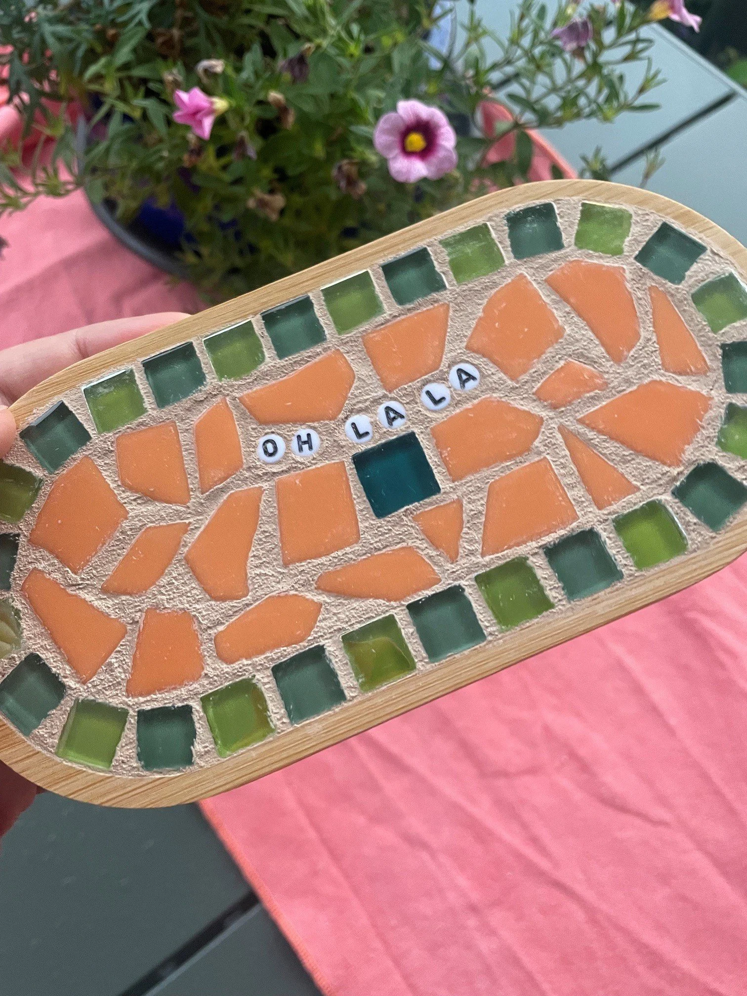 Eine Mosaikschale mit orange- und grünen Glasteilen, auf der die Buchstaben 'OH LA LA' zu sehen sind, gehalten in einer Hand vor einem Blumenpott mit pinken und lila Blumen.