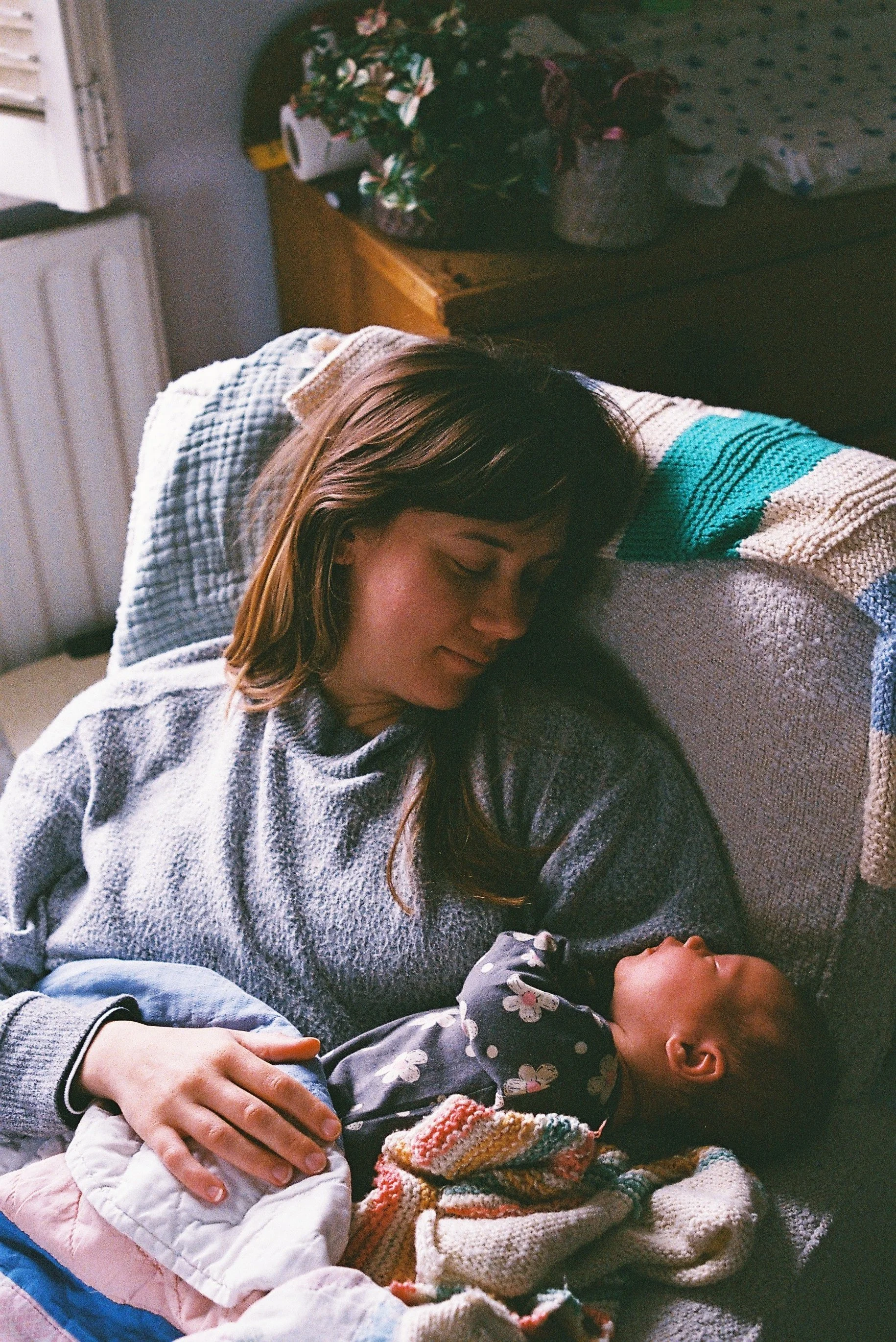 A woman with brown hair, wearing a grey sweater, is gently holding and lying with a baby girl who is dressed in a black floral outfit and wrapped in a multi-colored knitted blanket. They are resting on a couch indoors.
