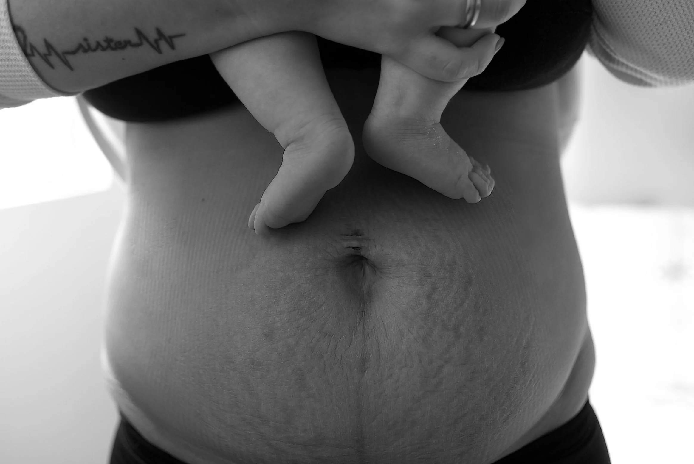 Close-up of a person's hand with a tattoo on the forearm, holding a foot with curled toes, and the person's torso visible in black and white. The person's torso is stretched to indicate postnatal recovery.