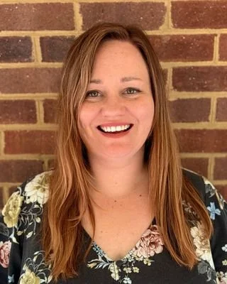 A woman with long, light brown hair, smiling, standing in front of a brick wall, wearing a floral top.