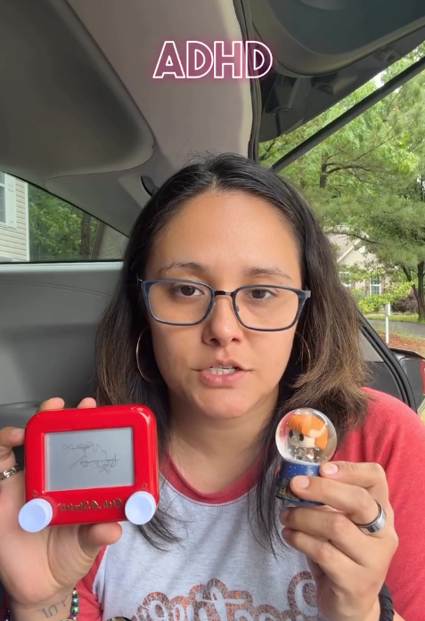 Woman with glasses and hoop earrings sitting in a car, holding a red Nintendo Game Boy and a snow globe with orange and white objects inside, with the word 'ADHD' at the top of the image.
