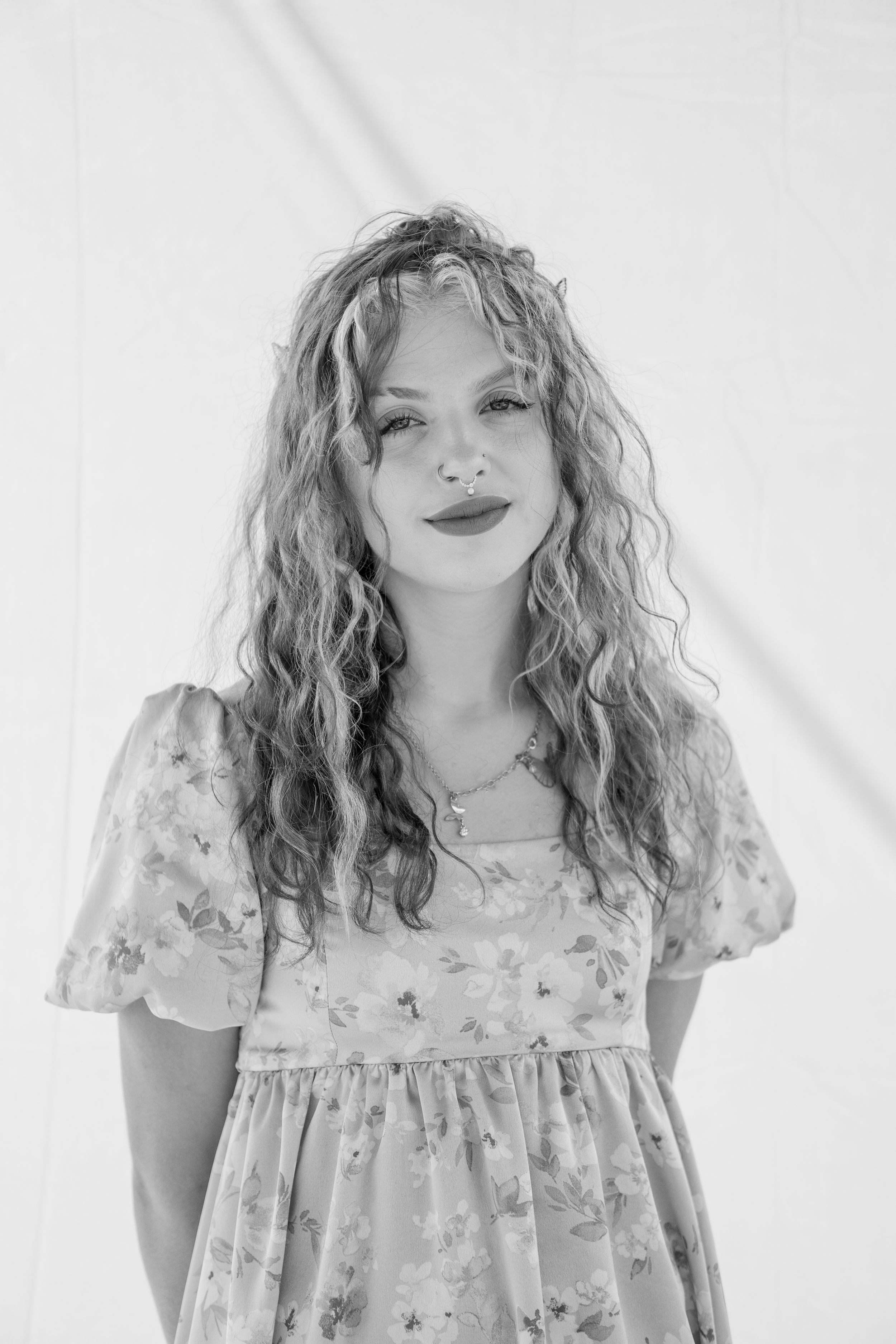 Black and white portrait of a young woman with curly hair, wearing a floral dress and layered necklaces, standing against a plain background.