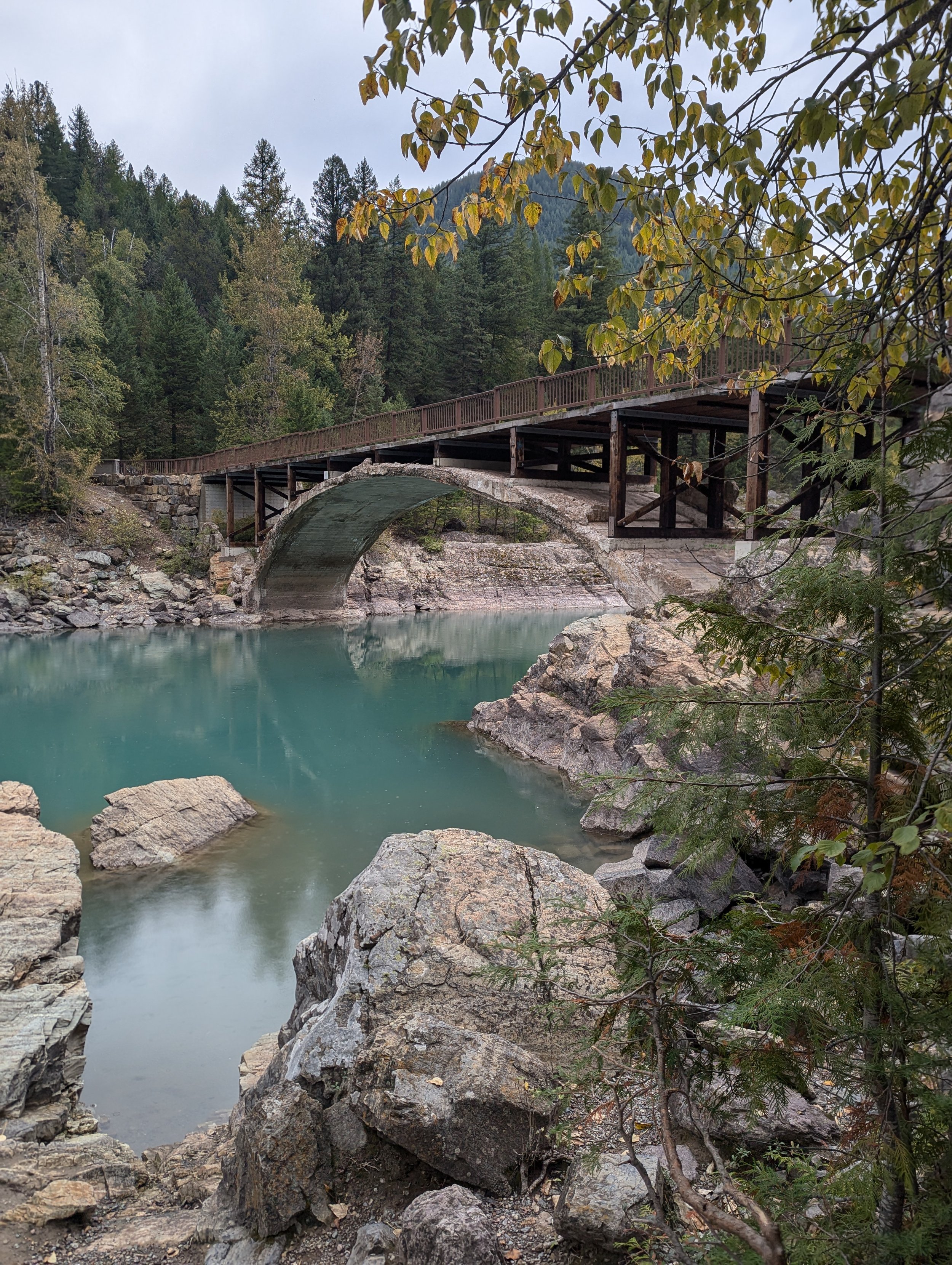 A wooden bridge over a rocky river in a forested mountain area. Green trees and hills in the background, overcast sky.