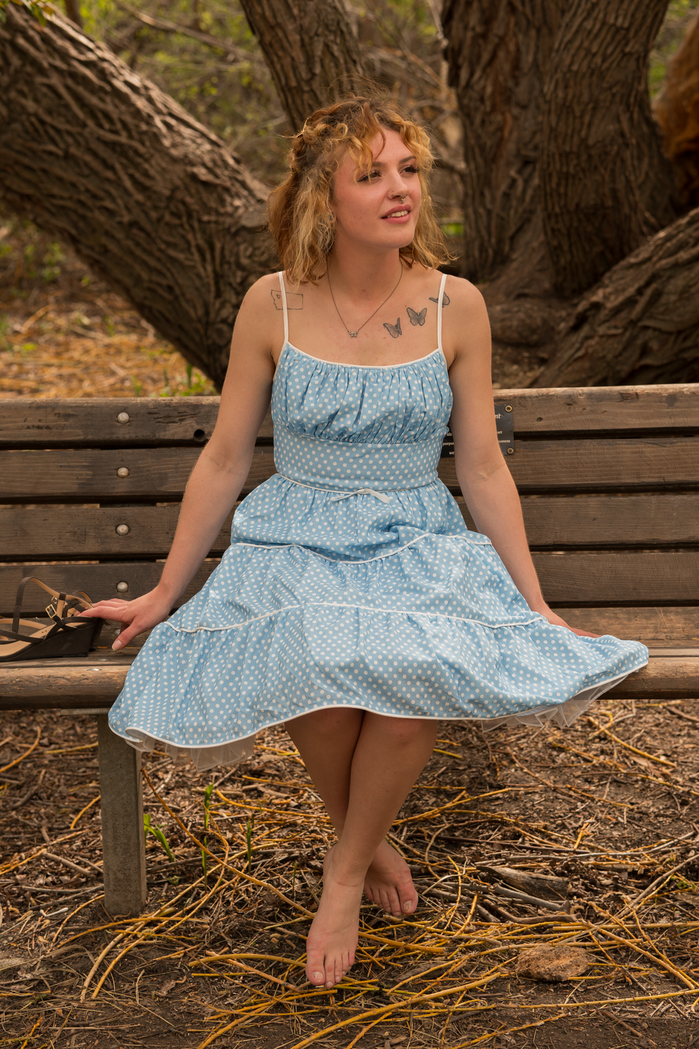 A young woman with curly blonde hair sitting barefoot on a park bench. She wears a blue polka dot dress and has tattoos of butterflies and a square on her chest. She is outdoors among large trees.