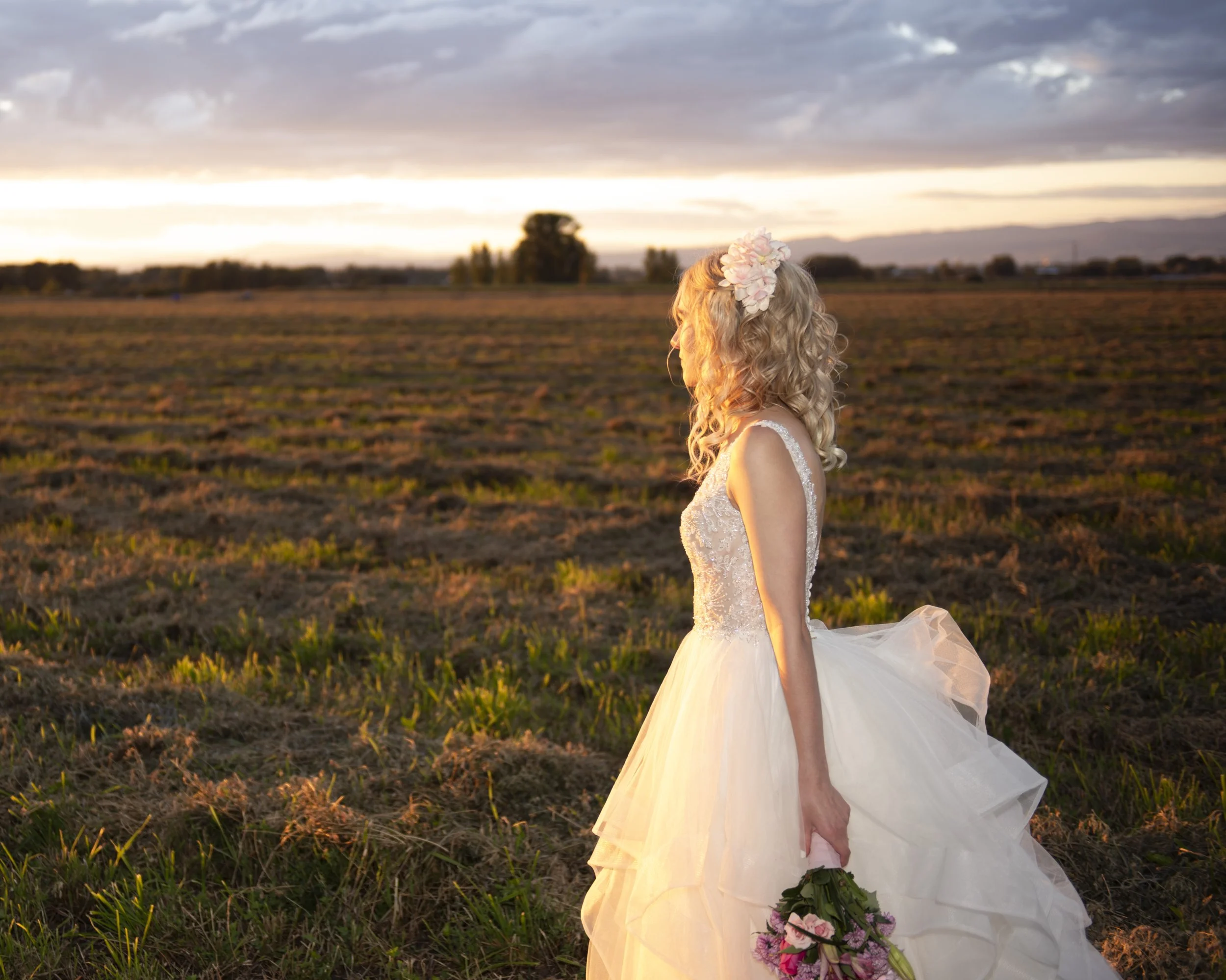 A woman in a white wedding dress holding a bouquet of pink roses and wearing a floral headband, standing in a plowed field at sunset with a cloudy sky in the background.
