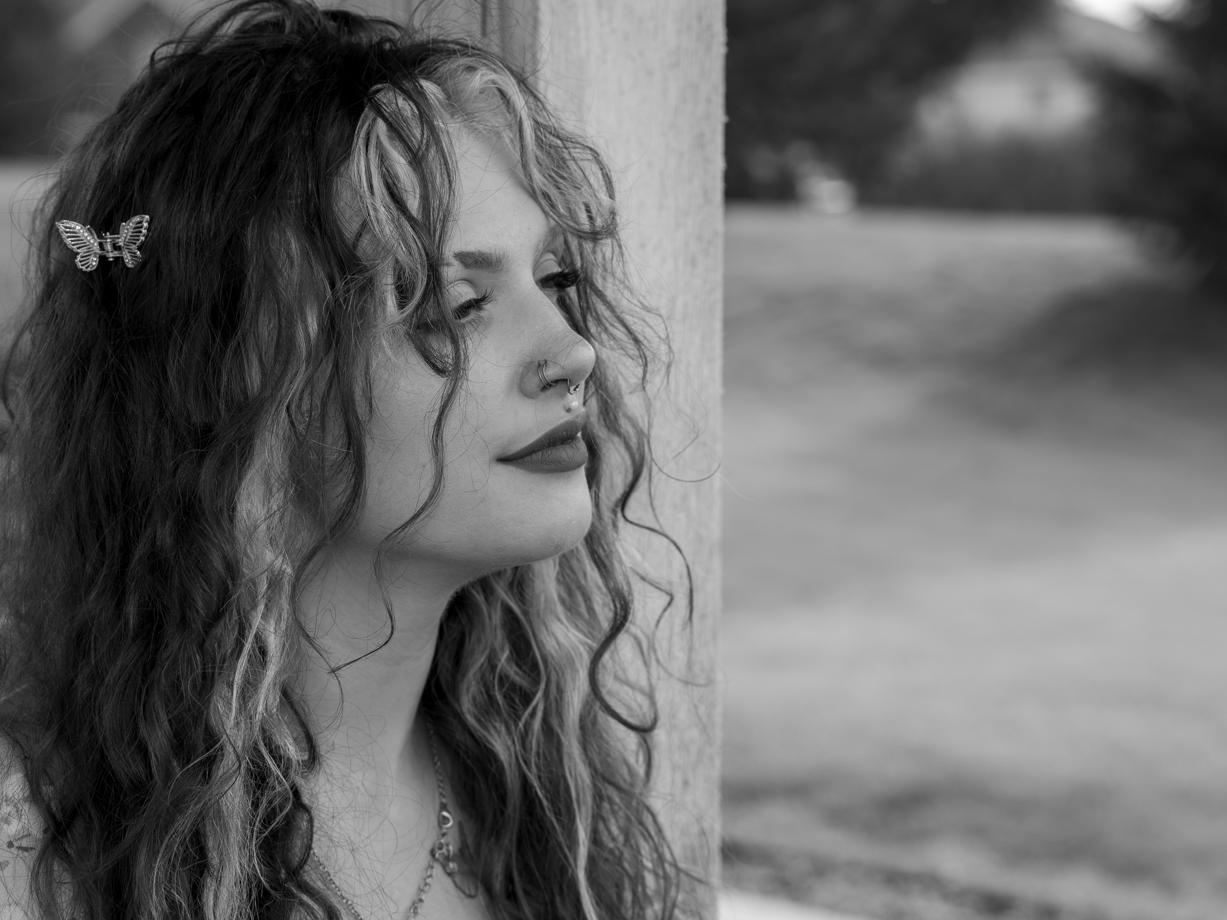 A black and white photo of a woman with curly hair decorated with a butterfly hair clip, standing beside a wooden post outdoors.