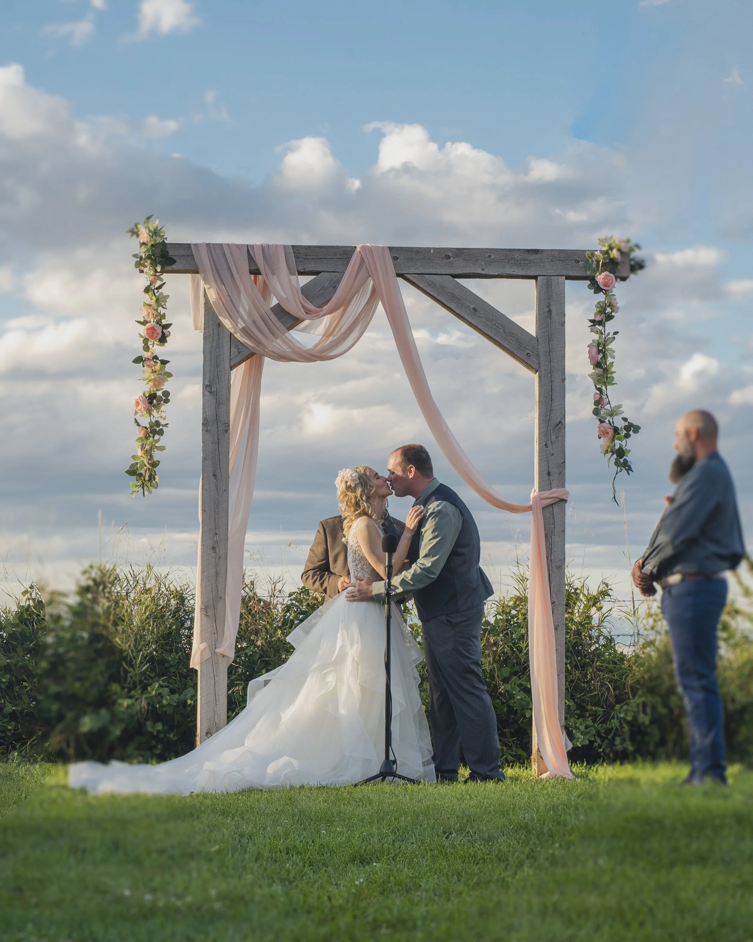 A couple is exchanging vows under a decorated wooden wedding arch outdoors, with a partly cloudy sky and grassy field in the background.
