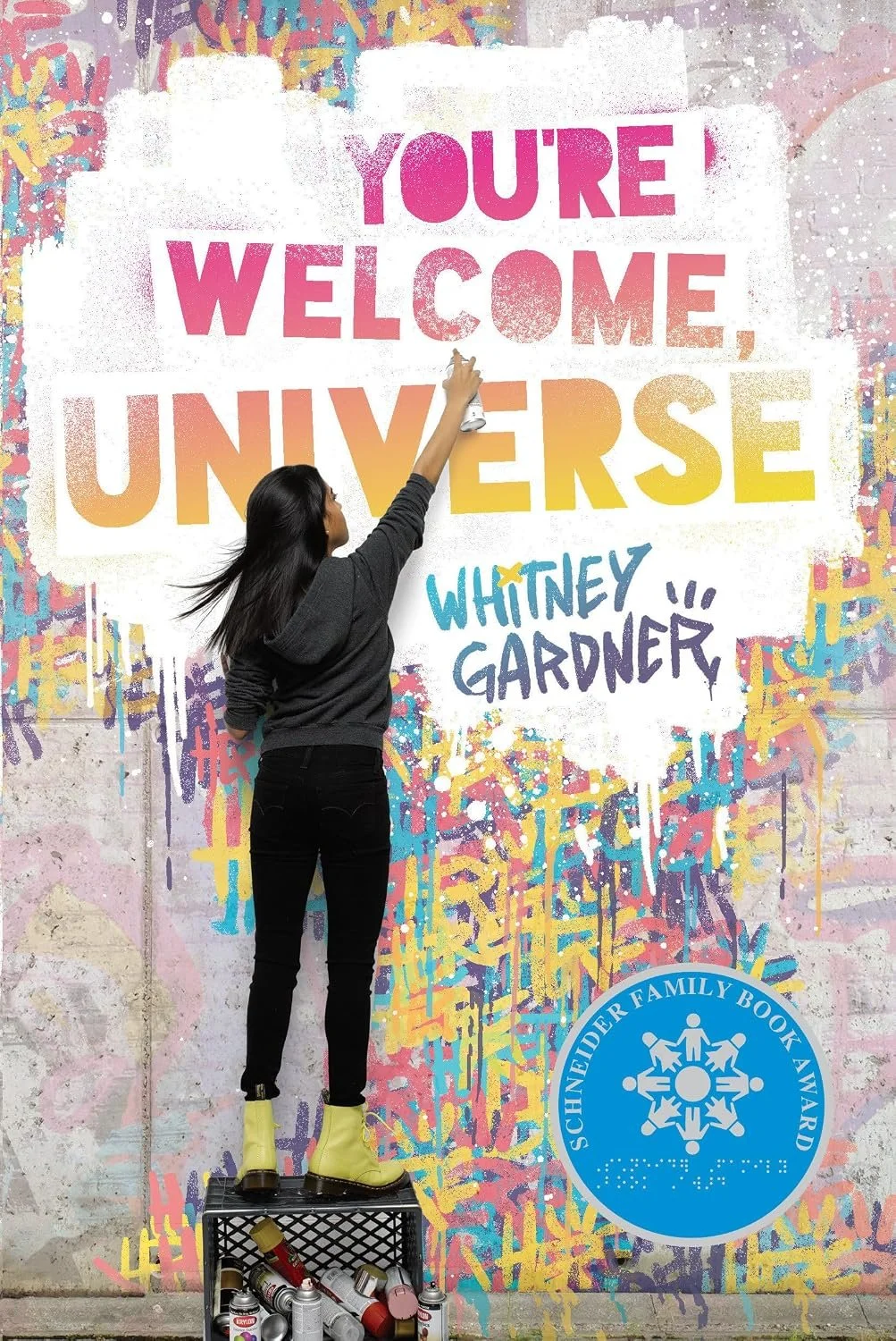 A woman spray painting a colorful mural on a wall that reads "You're welcome, universe" and is signed by Whitney Gardner, with a blue circular sticker referencing the Schneider Family Book Award. She is standing on a crate with spray paint cans.