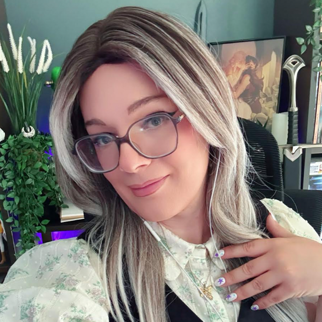 Mara Holmes (she/her) has long blonde hair, glasses, light makeup, and is wearing a pale yellow collared top and white earphones.