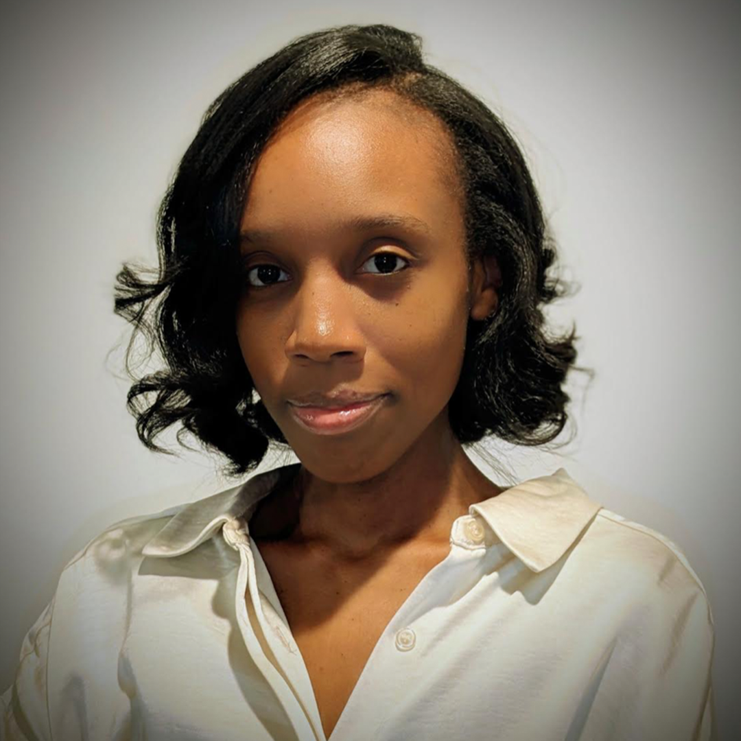Maia Wilson (she/her) has black curled hair, brown eyes, and is wearing a white collared top with the top few buttons left open. She is smiling in front of a grey background.