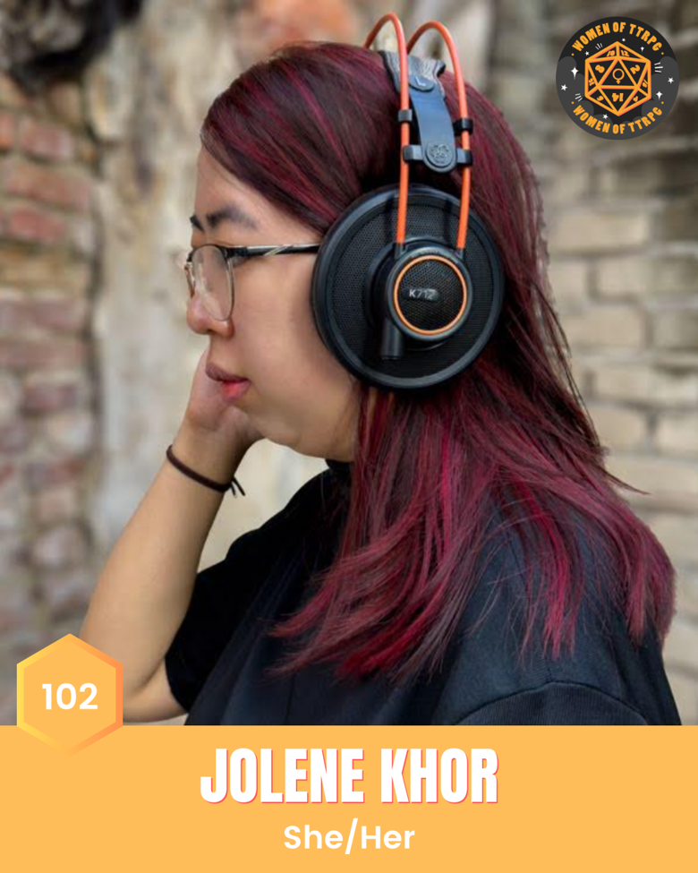 Jolene Khor (she/her) faces the side with long red hear, wearing a black tshirt, glasses, and headphones.